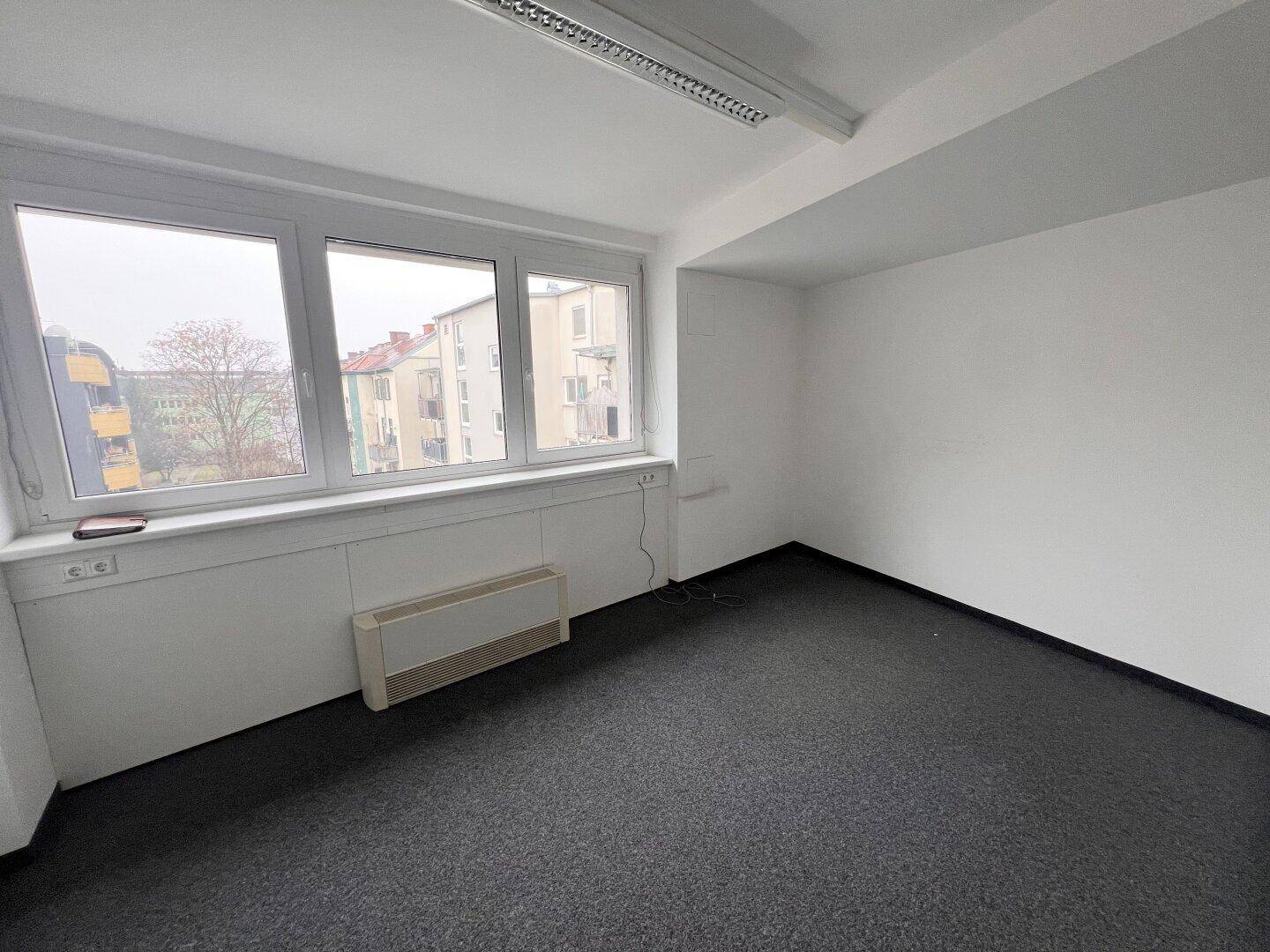 Leeres Büro mit Teppichboden, großen Fenstern und Blick auf die umliegenden Gebäude.