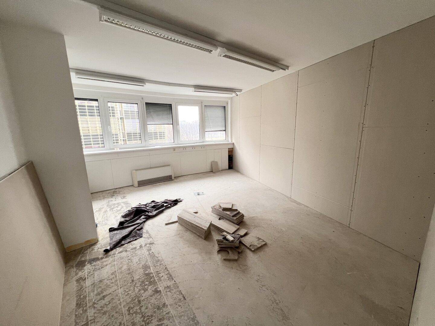 Großes Büro im Rohbau mit Trockenbauwänden, unfertigem Boden und Fensterfront mit Jalousien.