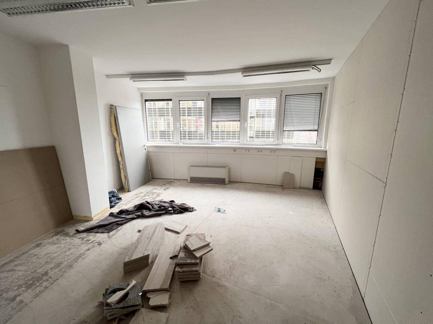 Geräumiges Büro im Rohbau mit Trockenbauwänden, unfertigem Boden und breiter Fensterfront mit Jalousien.