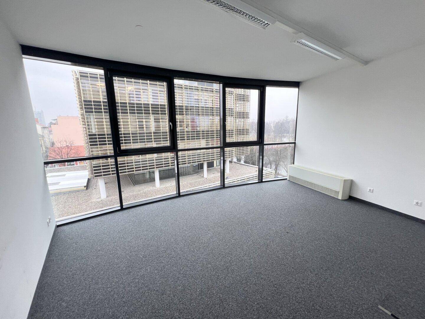 Leeres Büro mit großen Fenstern, Teppichboden und Blick auf die moderne Gebäudefassade.