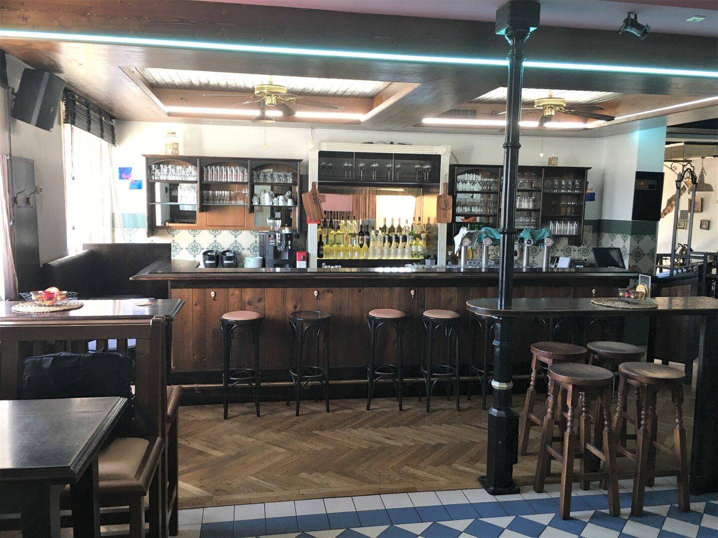 Innenansicht einer Bar mit Holzelementen, Theke, Hockern und einem gemusterten Fliesenboden.