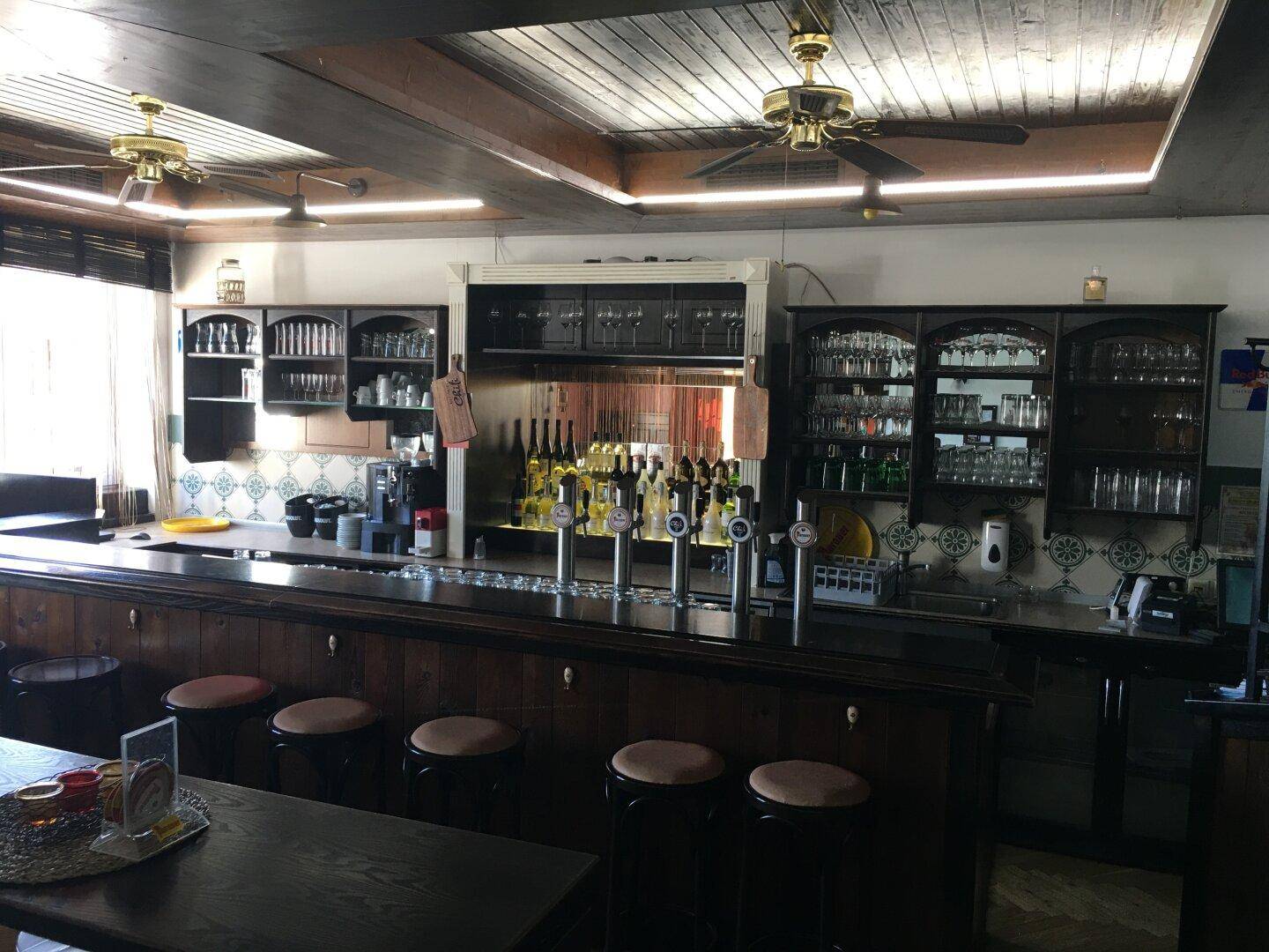 Geräumige Bar mit Holztheke, Deckenventilatoren und einer Auswahl an Getränken und Gläsern.