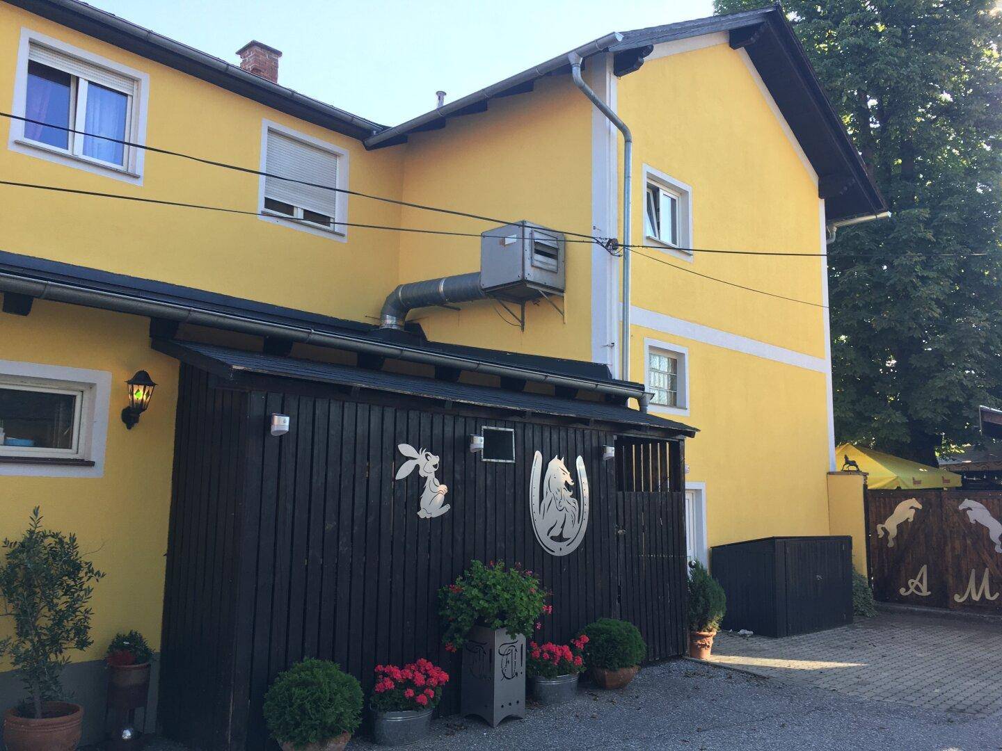 Gelbes Gebäude mit dunklem Holzverschlag, externer Lüftungsanlage und dekorativen Elementen an der Fassade.
