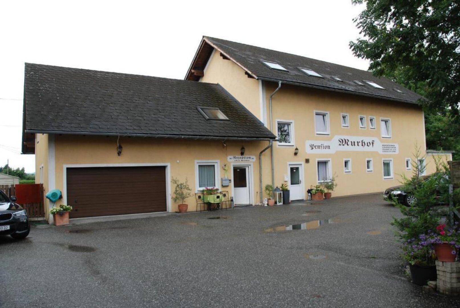 Seitenansicht eines gelben Gebäudes mit Garage, Eingangsbereich und Parkplatz, umgeben von Bäumen.