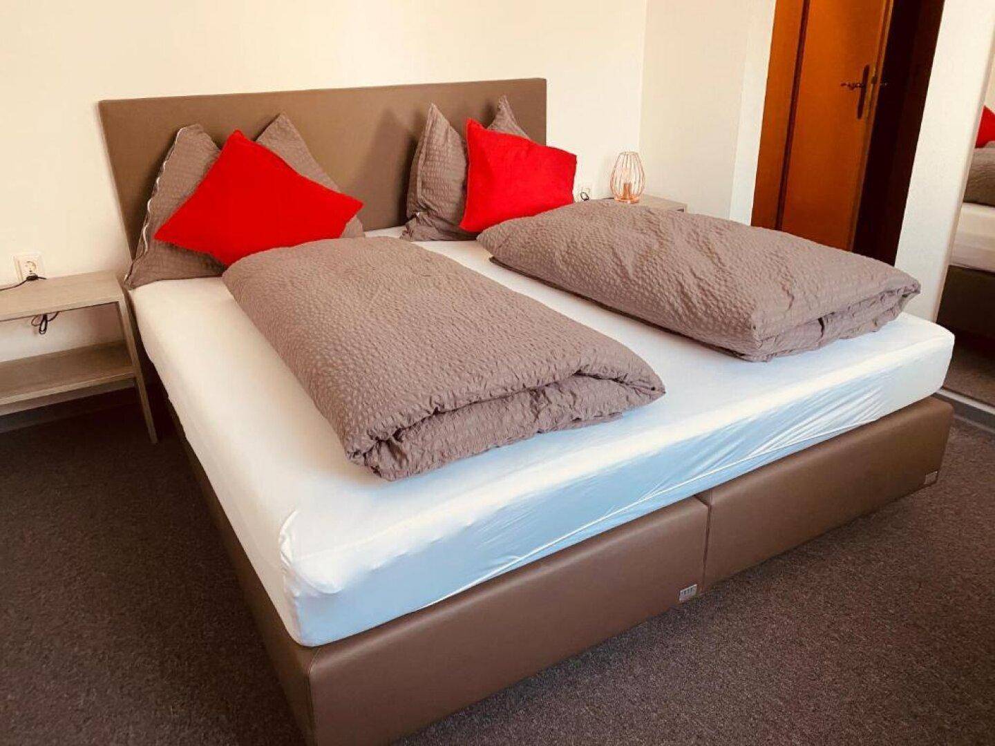 Gemütliches Schlafzimmer mit Doppelbett, Nachttischen und roten Kissen, in warmen Farbtönen gehalten.