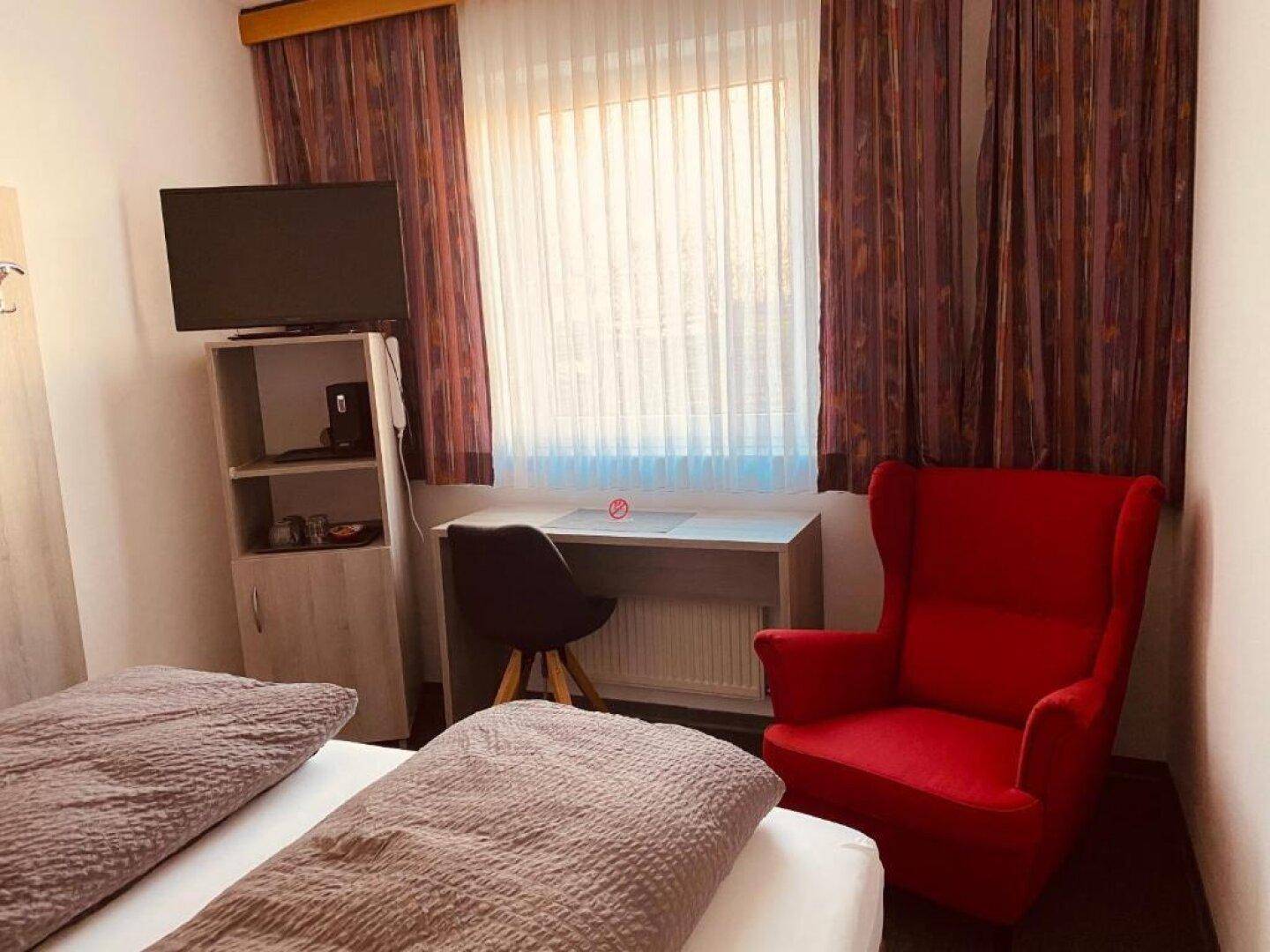 Schlafzimmer mit Einzelbetten, Schreibtisch, rotem Sessel und Fernseher am Fenster.