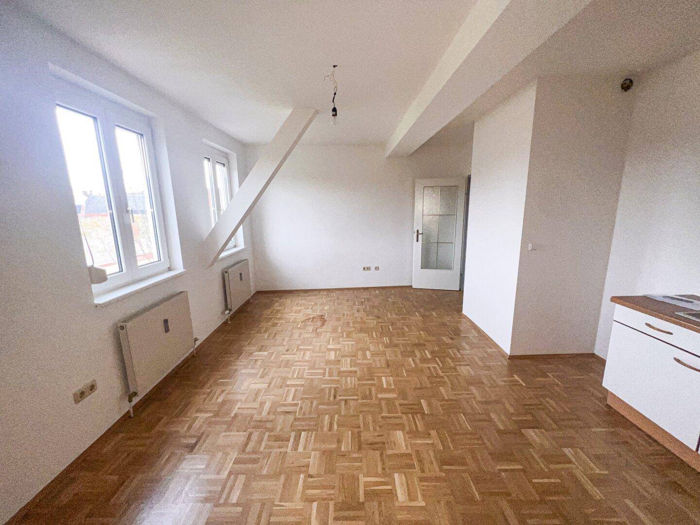 Geräumiger Wohnbereich mit Parkettboden, großen Fenstern und Zugang zu einem weiteren Raum.