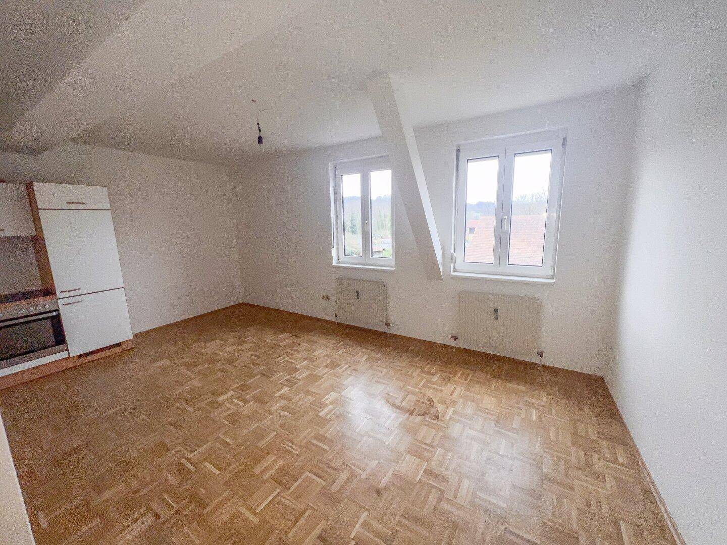 Offener Wohnbereich mit Parkettboden, zwei Fenstern und Heizkörpern unter den Dachschrägen.