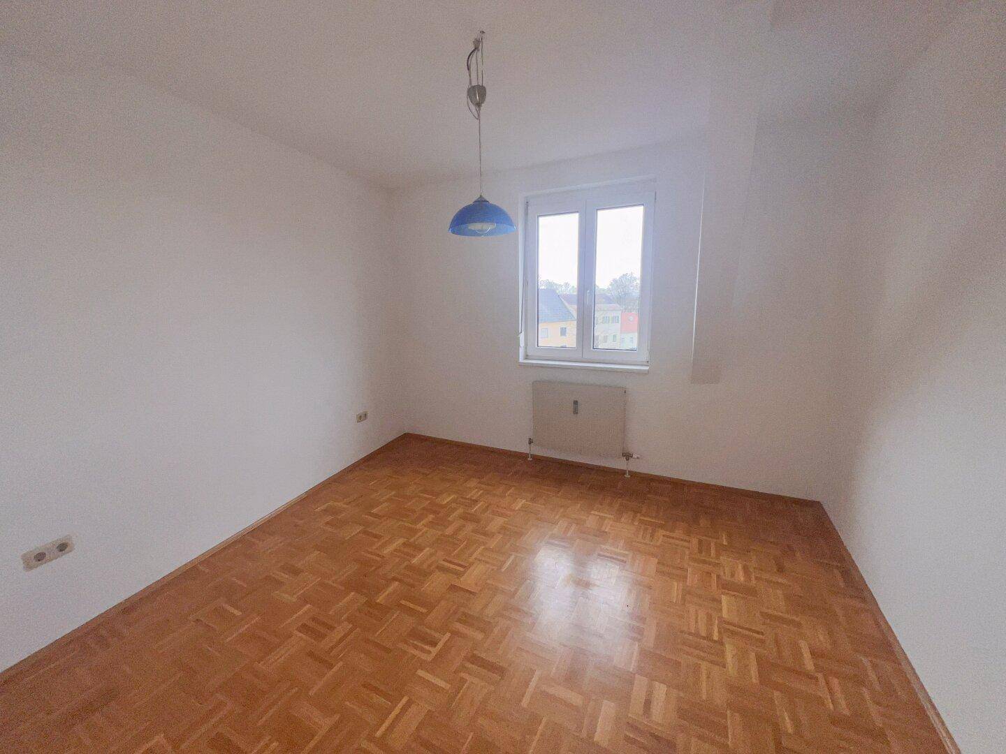 Helles Schlafzimmer mit Parkettboden, einem Fenster und einer blauen Deckenlampe.