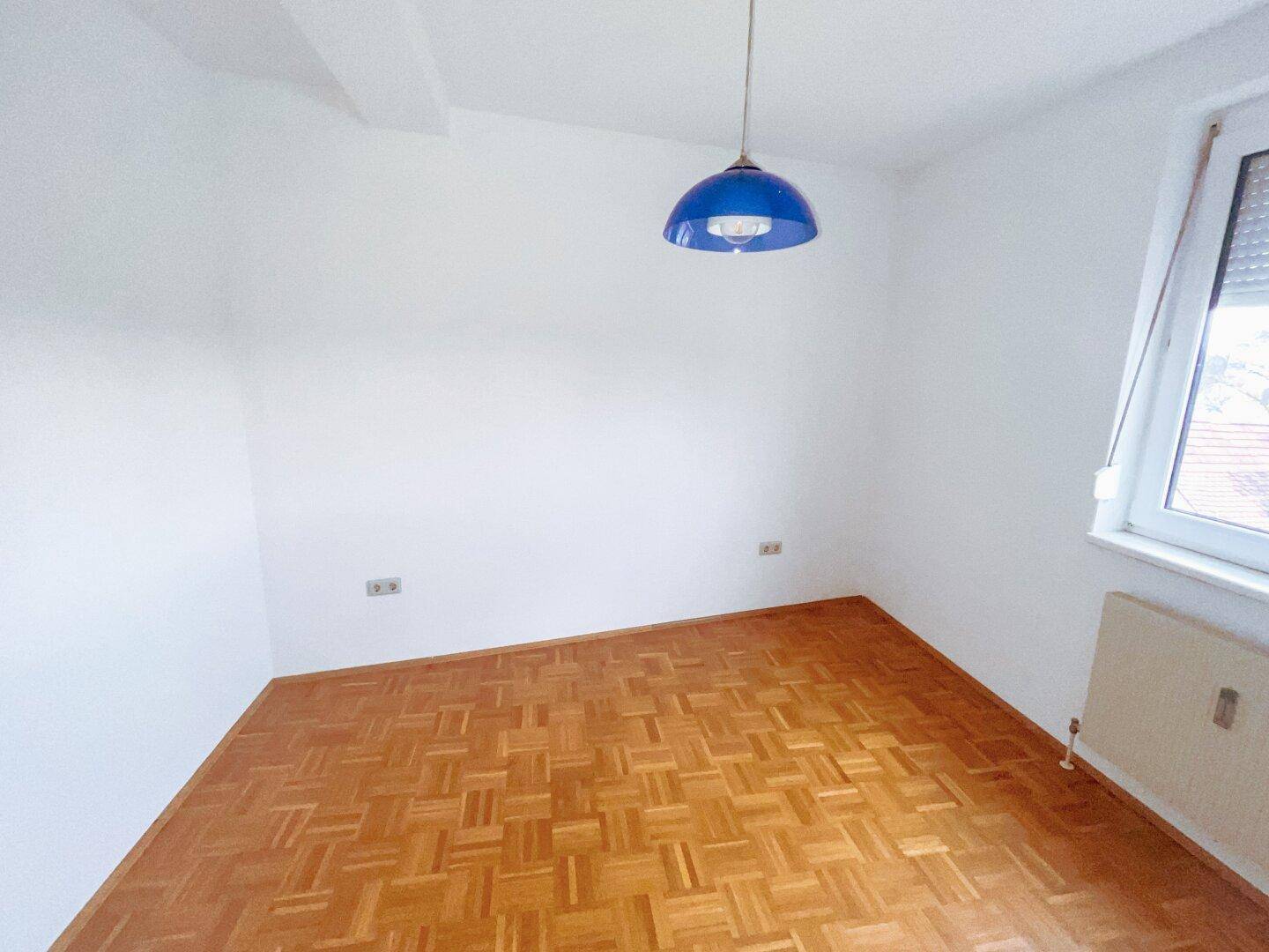 Schlafzimmer mit Parkettboden, weiß gestrichenen Wänden und einem Fenster mit Rollladen.