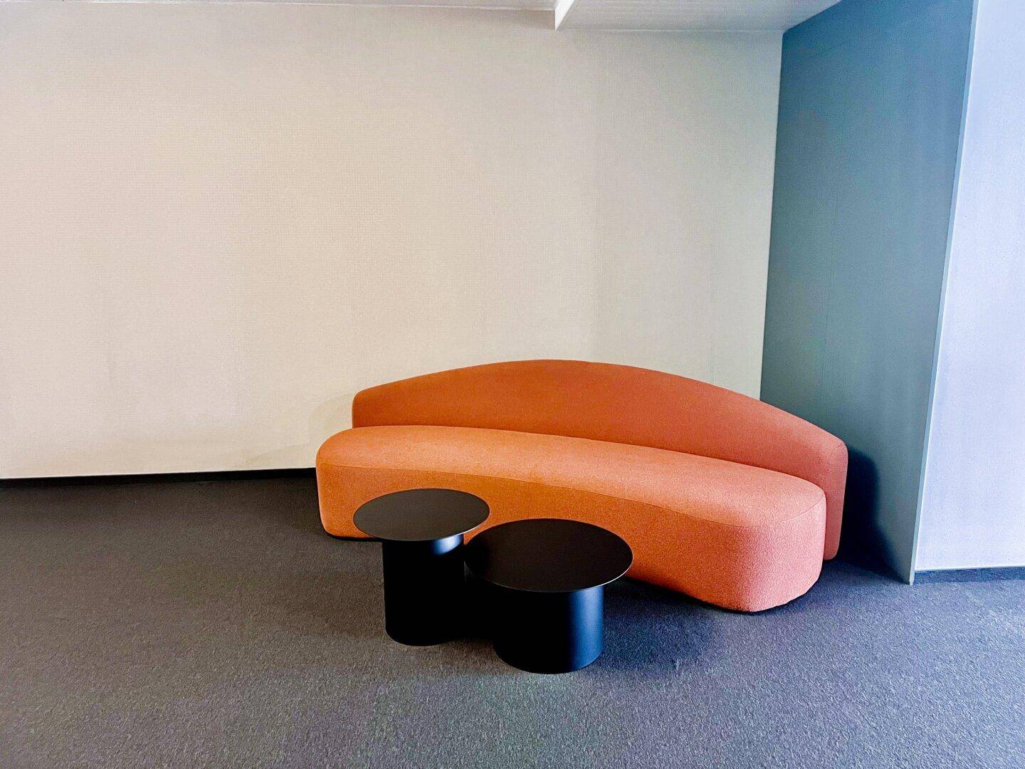Moderner Lounge-Bereich mit einem geschwungenen orangefarbenen Sofa und zwei runden schwarzen Beistelltischen.