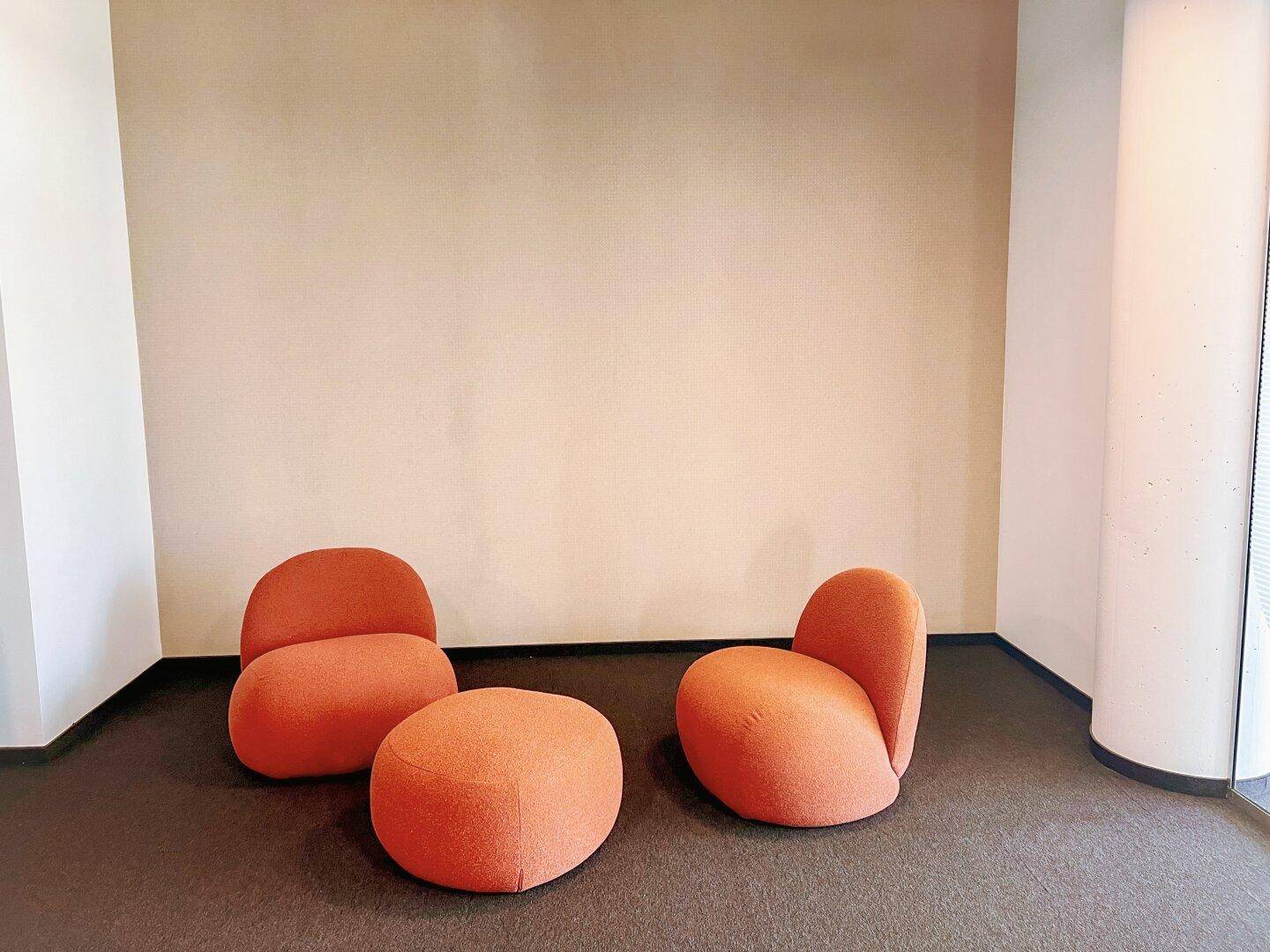 Gemütliche Lounge-Ecke mit drei orangefarbenen, runden Sitzmöbeln auf dunklem Teppichboden.