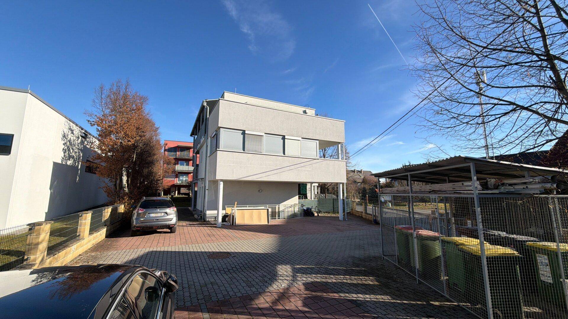 Rückansicht des Gebäudes mit gepflastertem Parkplatz und Mülltonnen unter klarem Himmel.