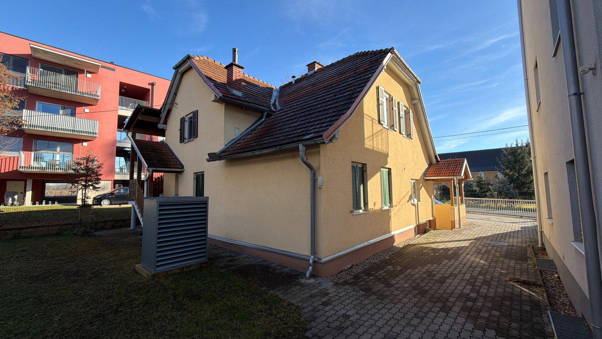 Seitenansicht des gelben Hauses mit Ziegeldach und einer Wärmepumpe im Gartenbereich.