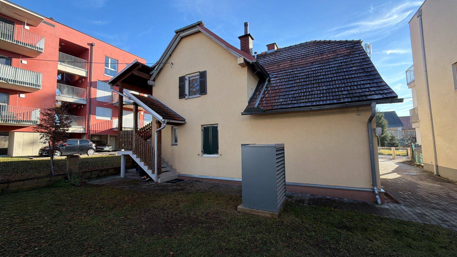 Rückseite des gelben Hauses mit Wärmepumpe, kleinem Garten und angrenzendem Neubau.