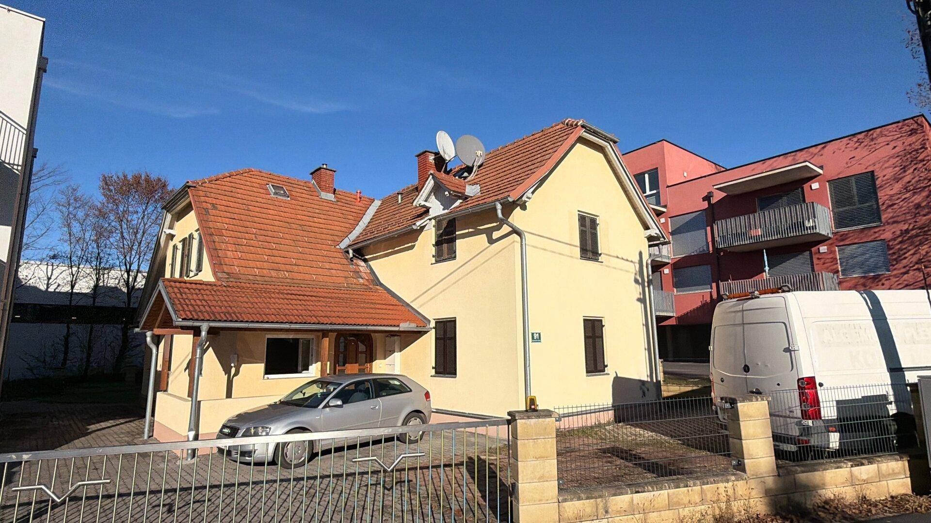 Älteres gelbes Wohnhaus mit Ziegeldach und Satellitenschüsseln, umgeben von moderneren Gebäuden.