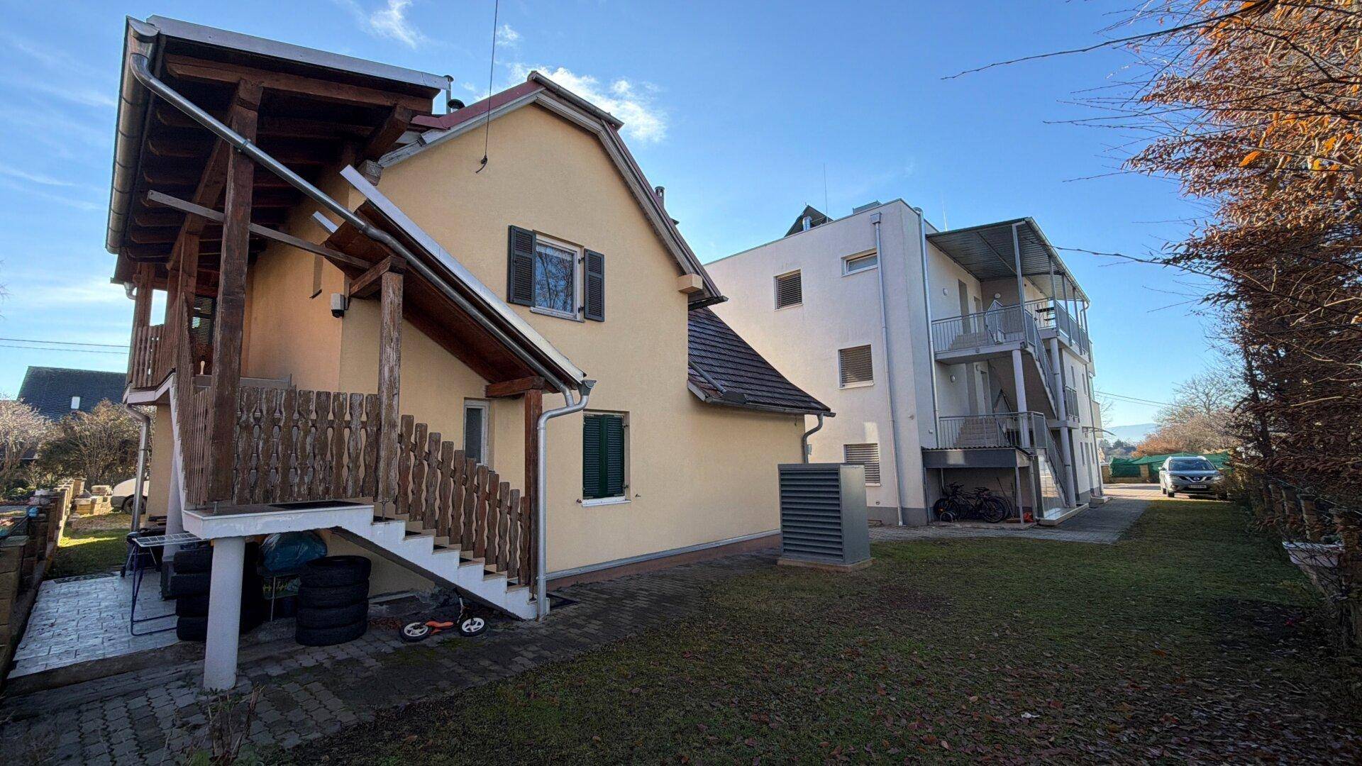 Gelbes Wohnhaus mit Außentreppe und Blick auf das moderne Nachbargebäude im Hintergrund.