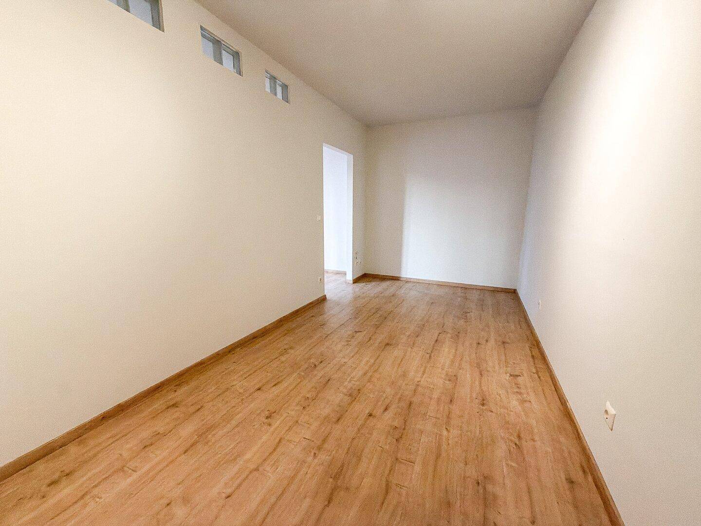 Geräumiger, leerer Wohnbereich mit hellem Laminatboden und weißen Wänden.