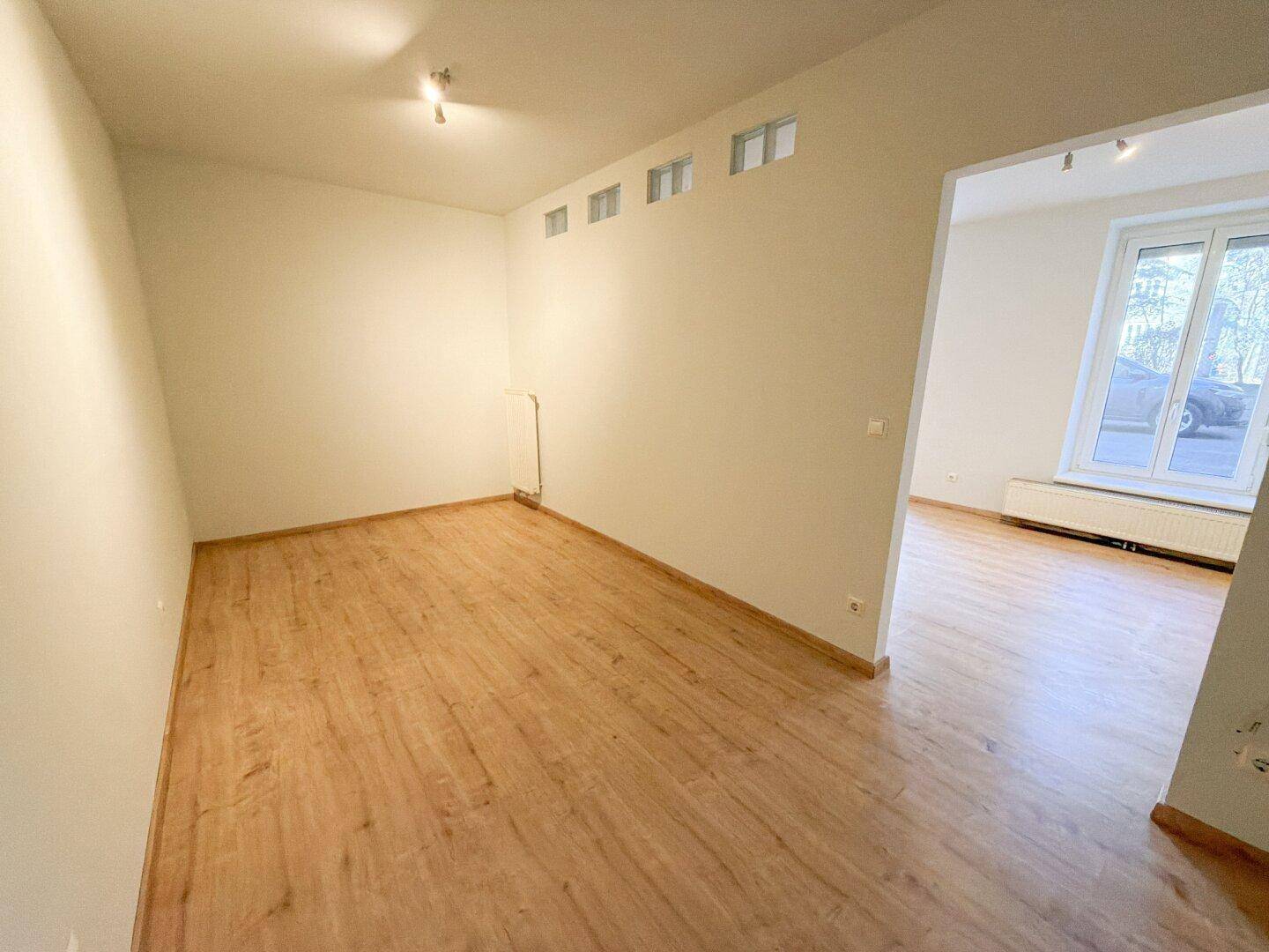 Heller Wohnbereich mit Laminatboden und Zugang zum Küchenbereich, gut beleuchtet durch ein großes Fenster.