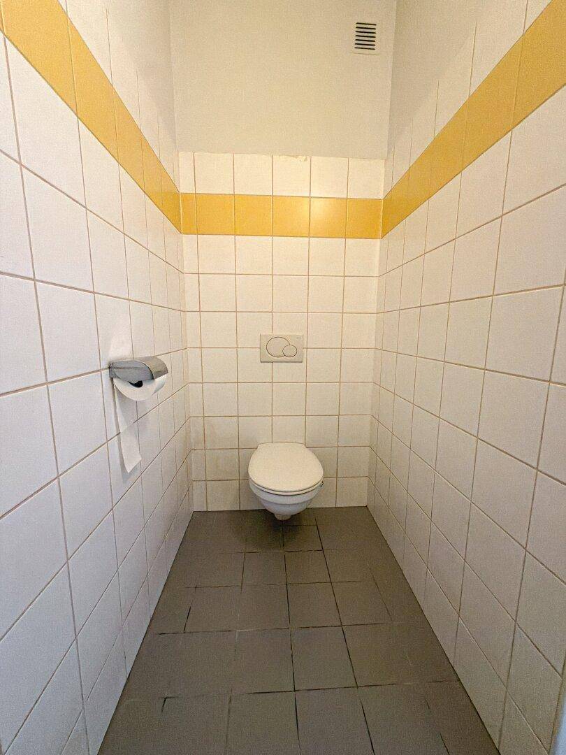 Separate Toilette mit weißen Fliesen, gelbem Akzentstreifen und dunklem Fliesenboden.