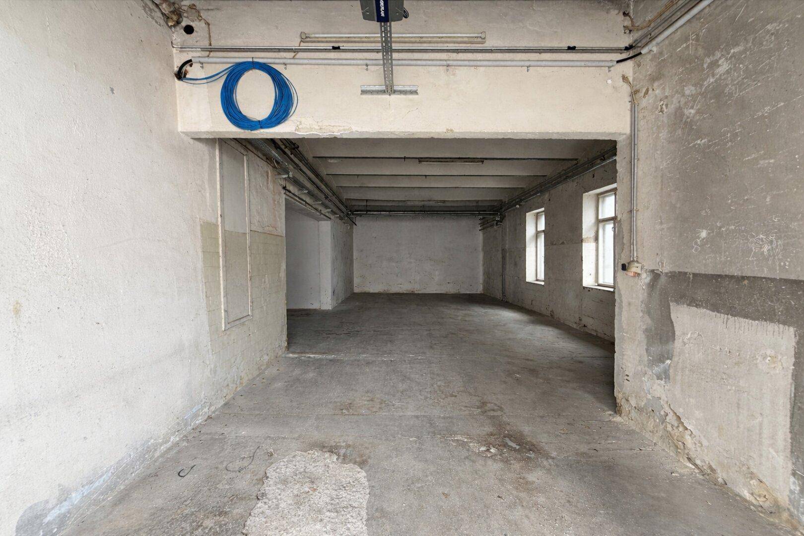 Langer, leerer Raum mit Betonboden, rauen Wänden und sichtbaren Kabeln und Leitungen an der Decke.