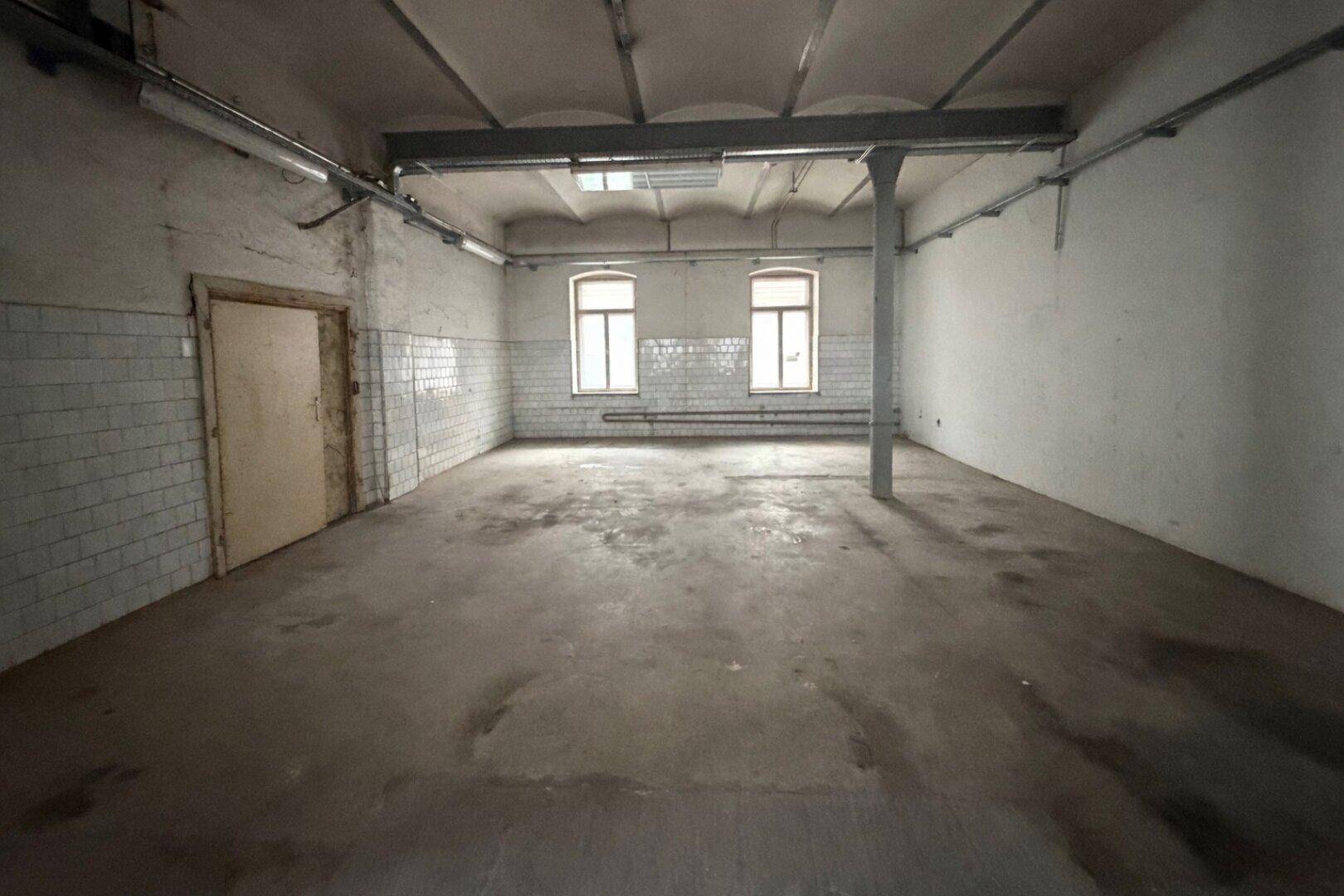 Geräumiger, leerer Gewerberaum mit Fliesen an den Wänden, Betonboden und zwei Fenstern.