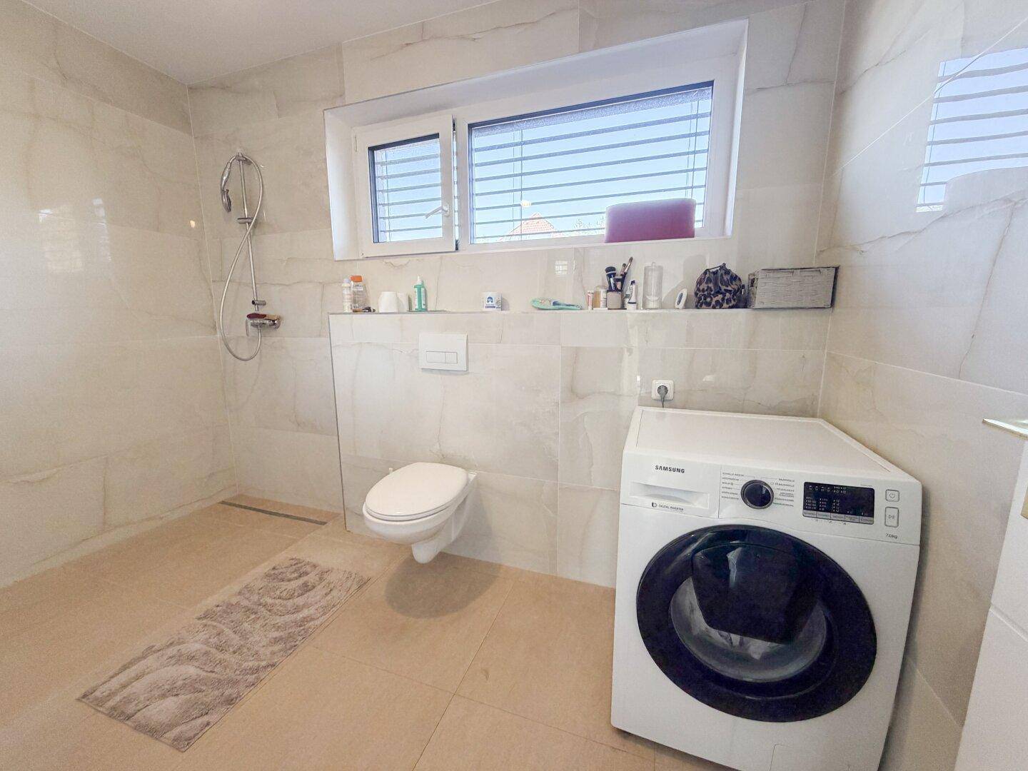 Helles Badezimmer mit Dusche, Wand-WC und Platz für eine Waschmaschine, gut beleuchtet.