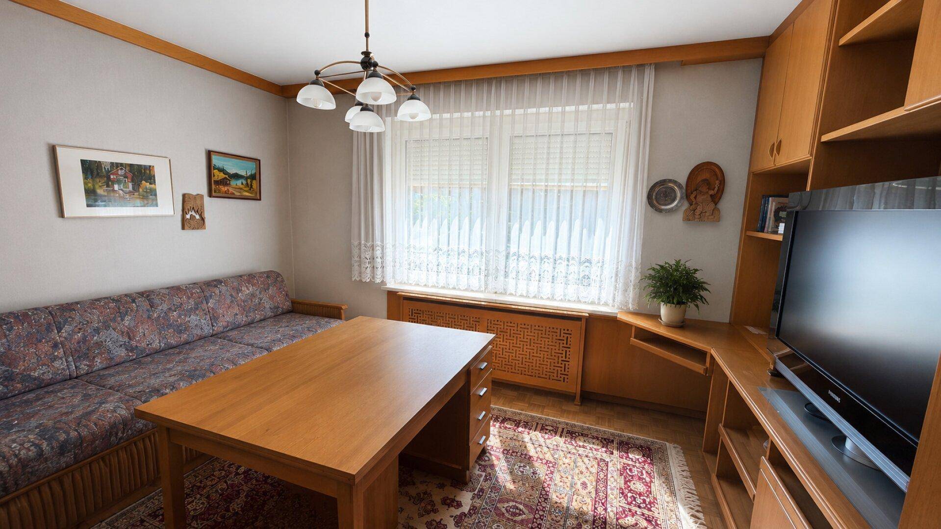 Kleines Arbeitszimmer mit Holzmöbeln, Teppich und Fenster mit Rolladen, funktional eingerichtet.