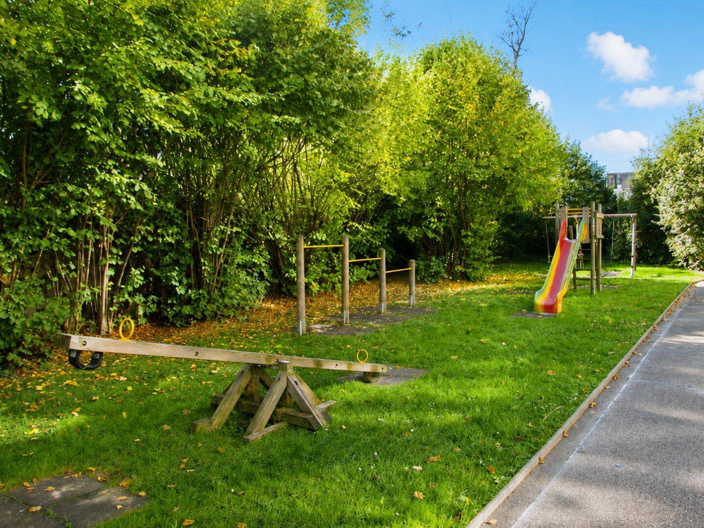 Grüner Spielplatz mit Wippe, Rutsche und Klettergerüst, umgeben von Bäumen.