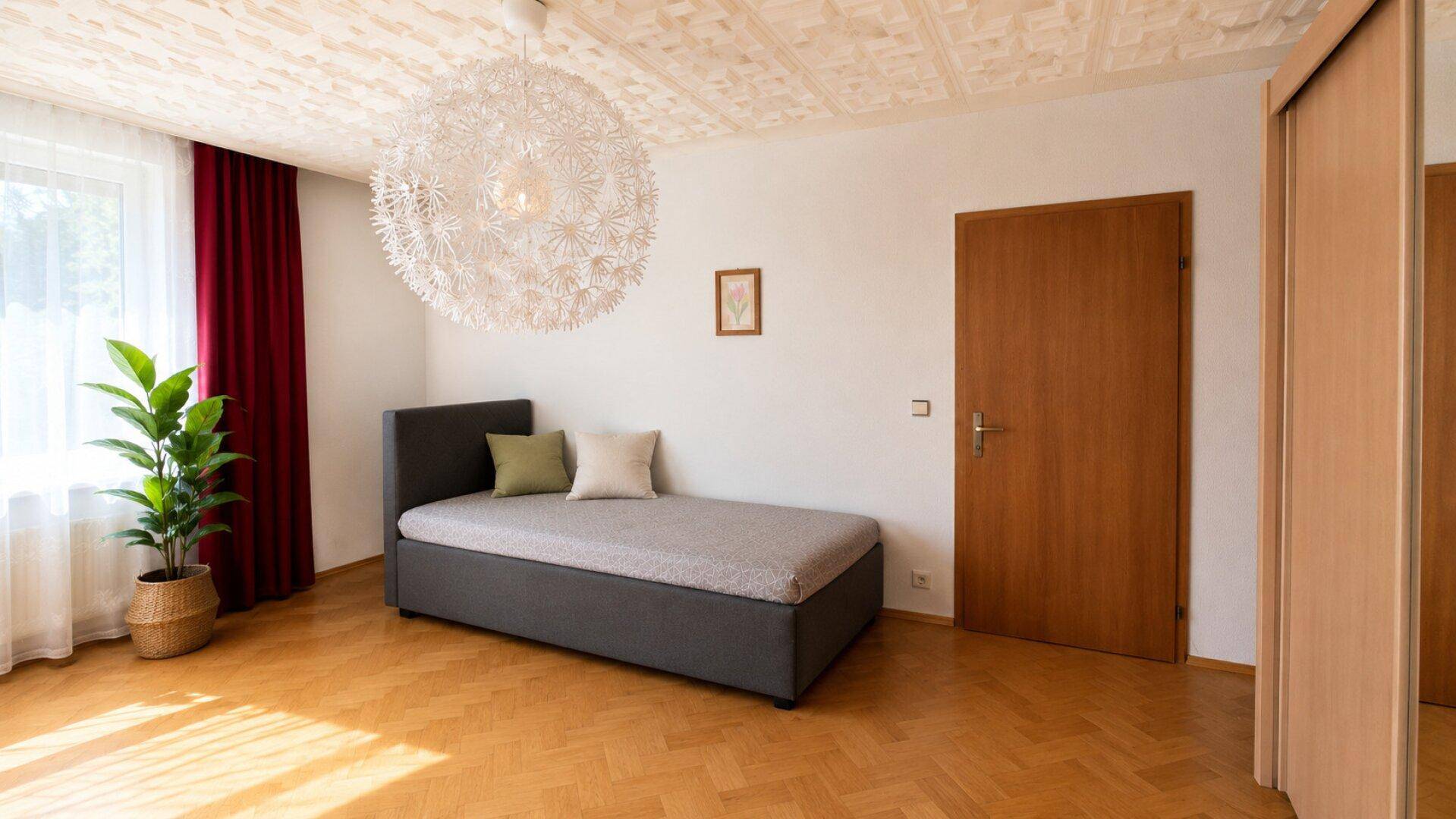 Helles Schlafzimmer mit Einzelbett, Holzschrank und dekorativer Deckenleuchte.