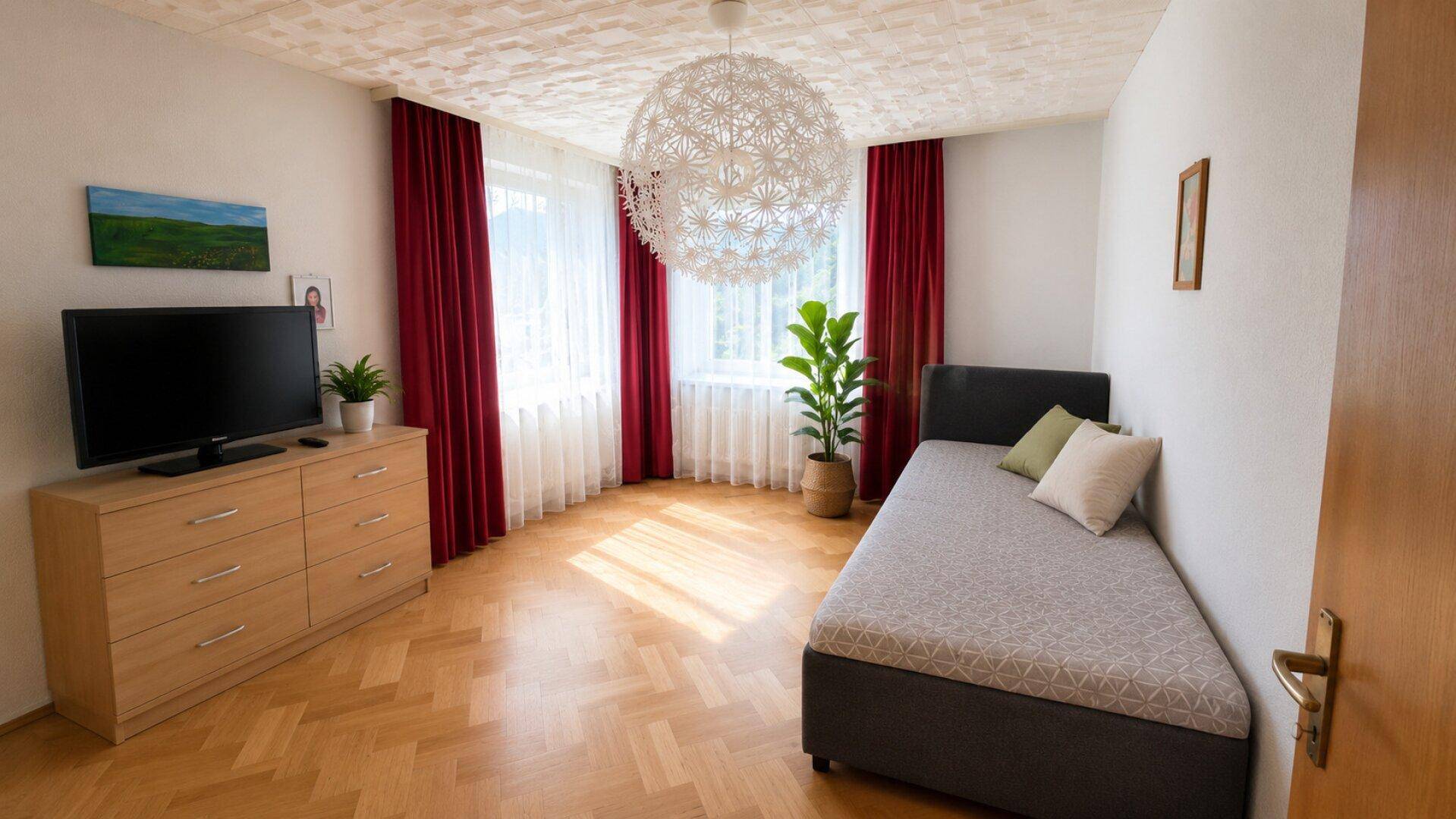 Schlafzimmer mit Einzelbett, Kommode und dekorativer Deckenleuchte, viel Tageslicht.