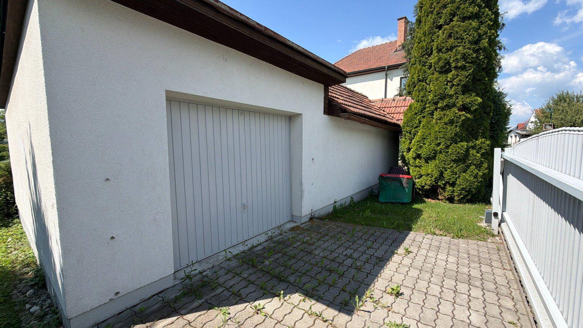 Weiße Garagenfassade mit gepflastertem Vorplatz und grüner Bepflanzung.