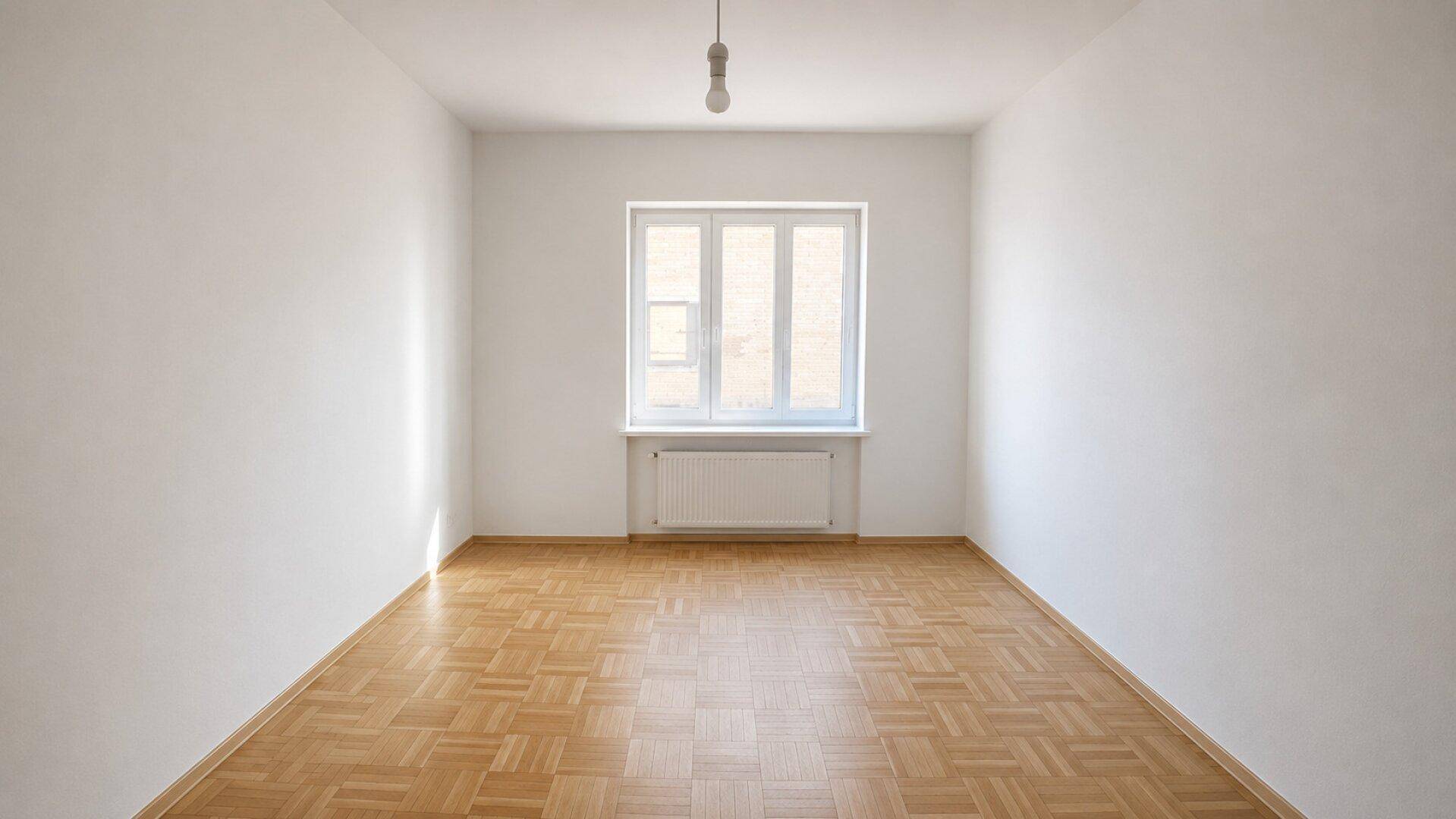 Leeres Wohnzimmer mit klassischem Parkettboden und großem Fenster für viel Tageslicht.