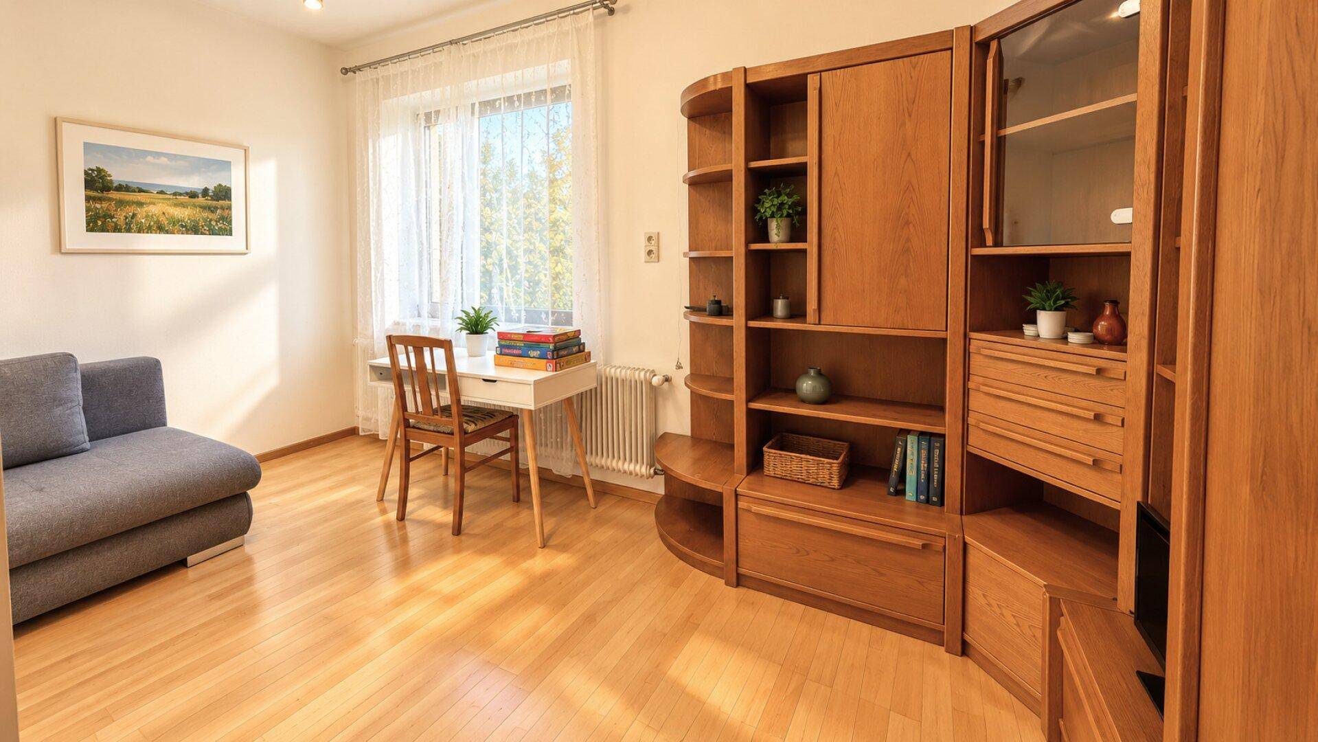 Heller Wohnbereich mit Holzboden, Schreibtisch am Fenster und einer großen Holzschrankwand.