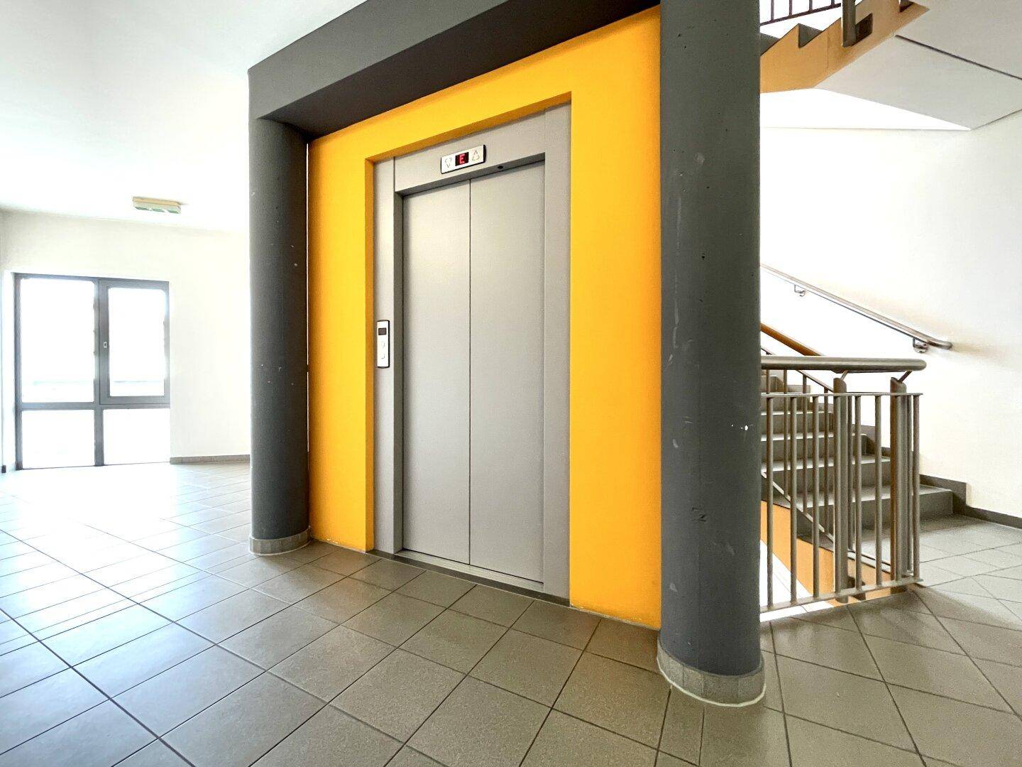 Eingangsbereich mit modernem Aufzug und Treppenhaus, gekennzeichnet durch gelbe Akzentwände und Fliesenboden.