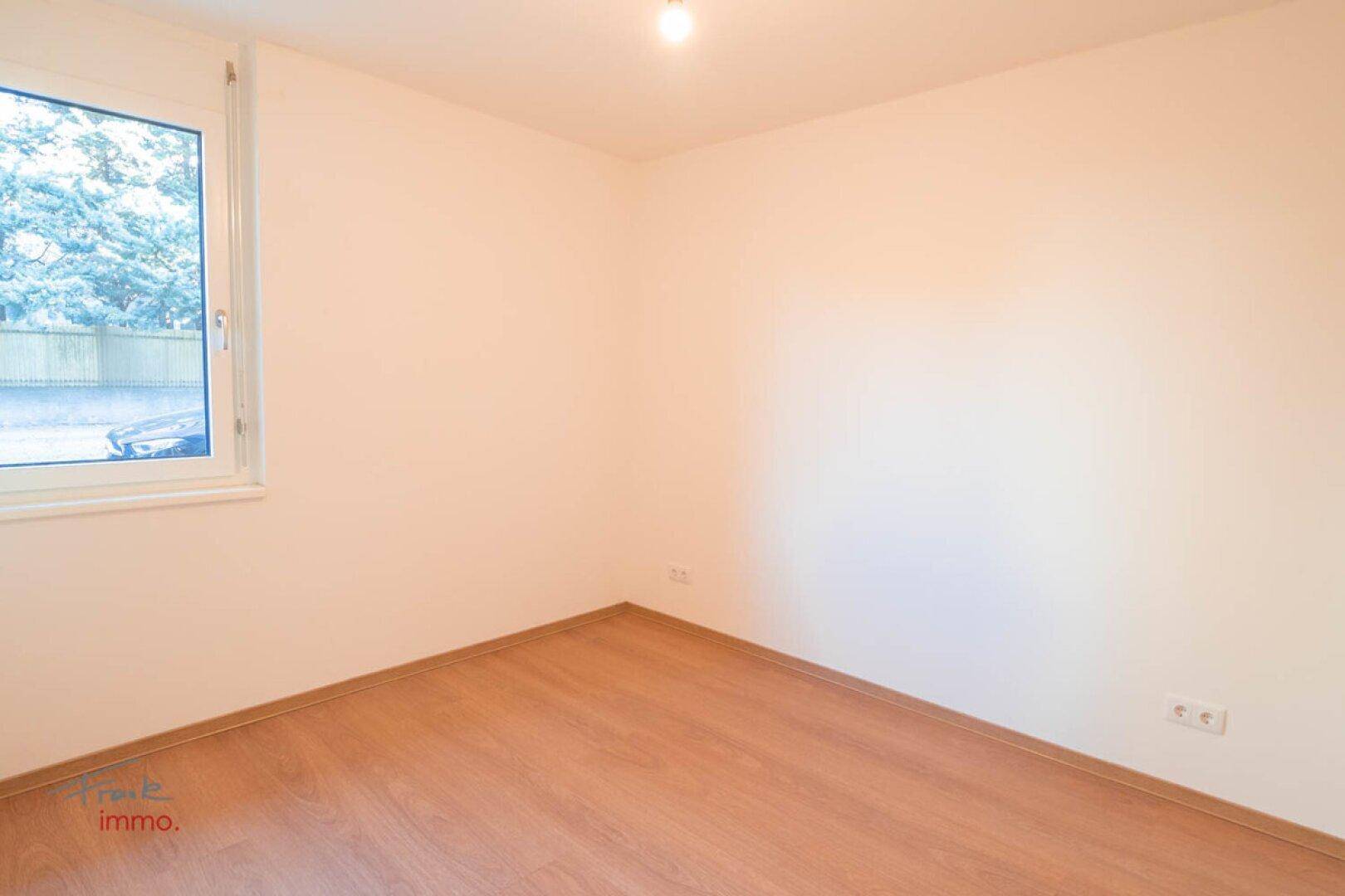 Leerer Raum mit hellem Holzboden und einem Fenster, das Tageslicht hereinlässt.