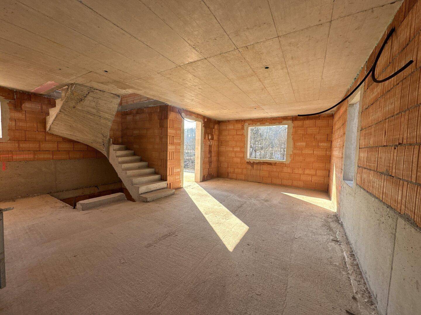 Innenraum eines Rohbaus mit Ziegelwänden, Betonboden und einer freitragenden Treppe.