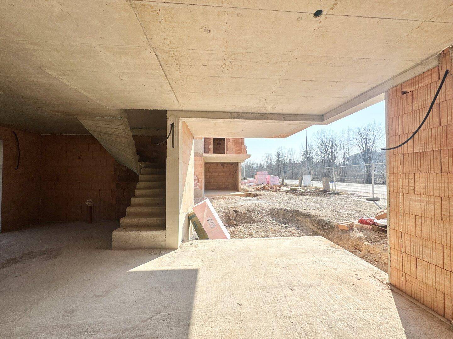 Unfertiger Innenraum eines Neubaus mit Betonboden und unverputzten Ziegelwänden, Blick nach draußen.