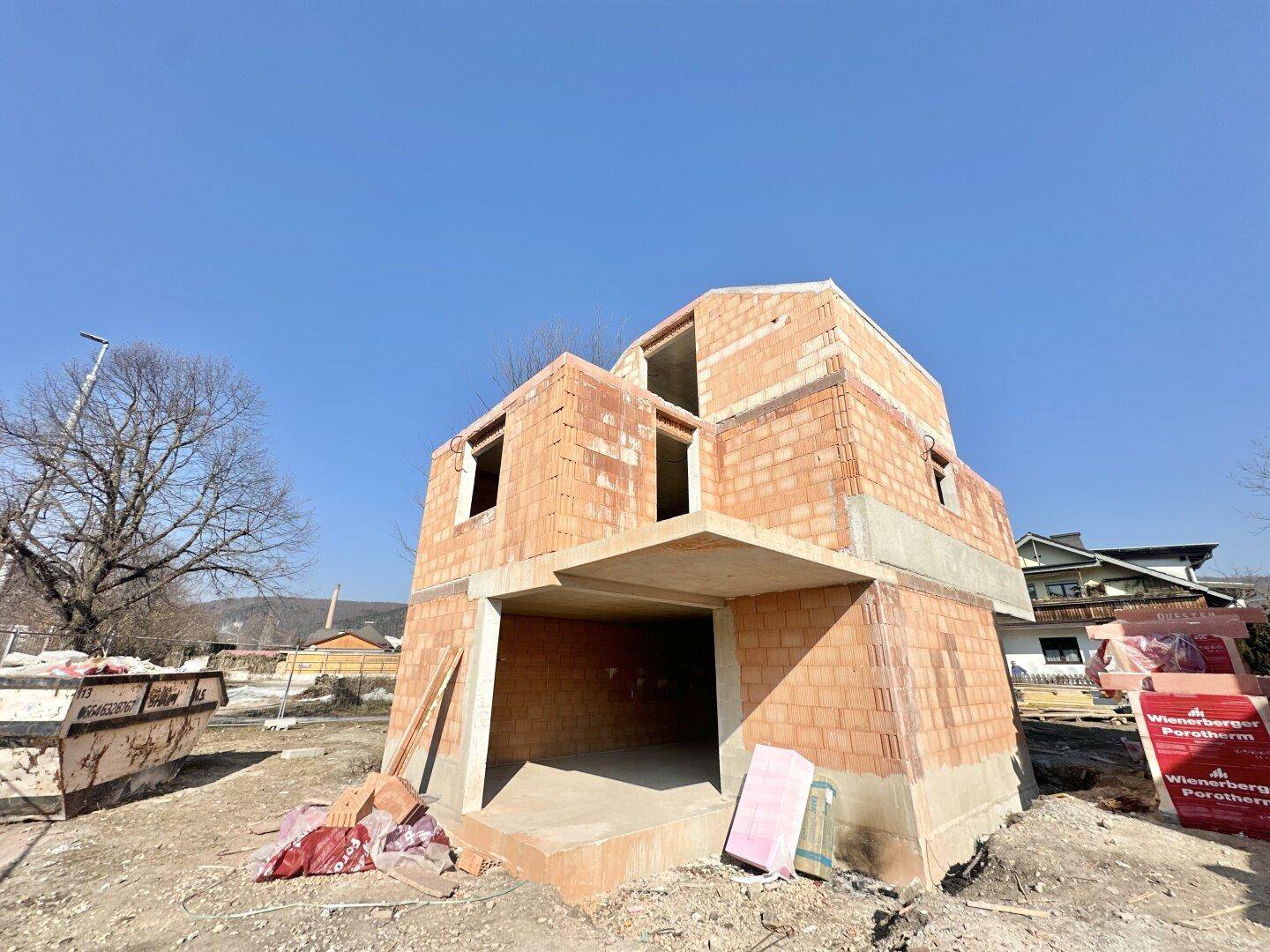 Rohbau eines Einfamilienhauses mit Ziegelwänden und Betonfundament unter strahlend blauem Himmel.