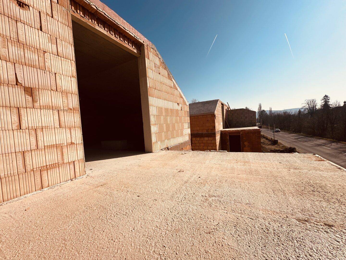 Rohbau eines Gebäudes mit unverputzten Ziegelwänden und einer angrenzenden Baustelle unter blauem Himmel.
