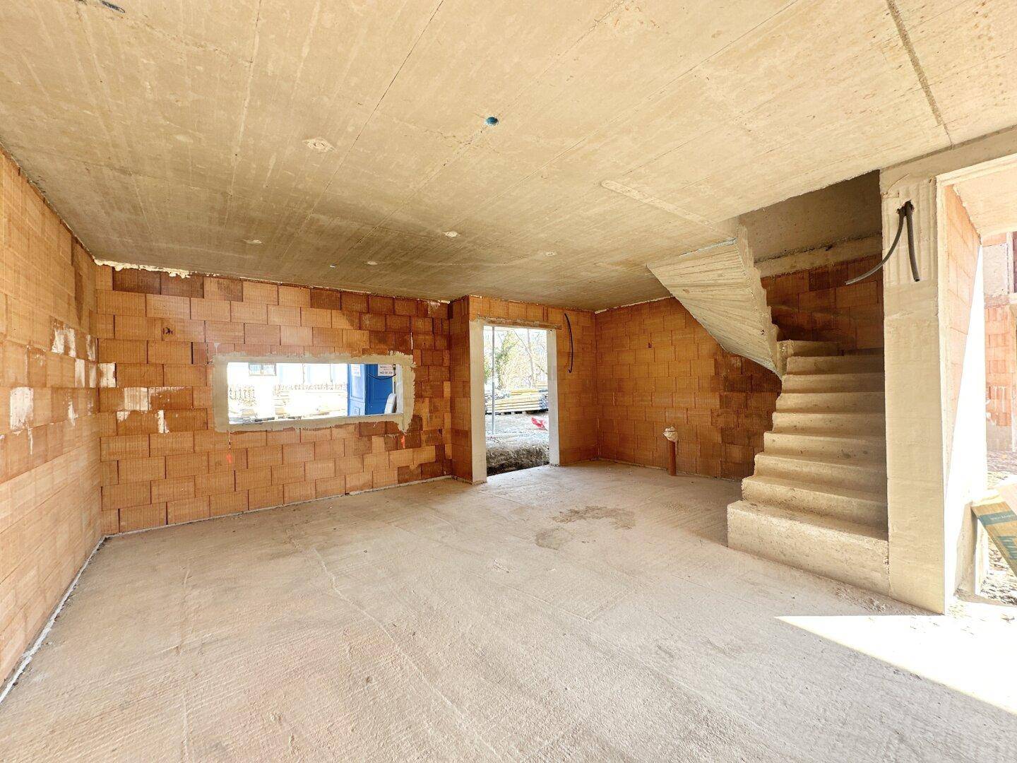 Geräumiger, unfertiger Innenraum mit Betonboden und einer freistehenden Betontreppe, ideal für den Wohnbereich.
