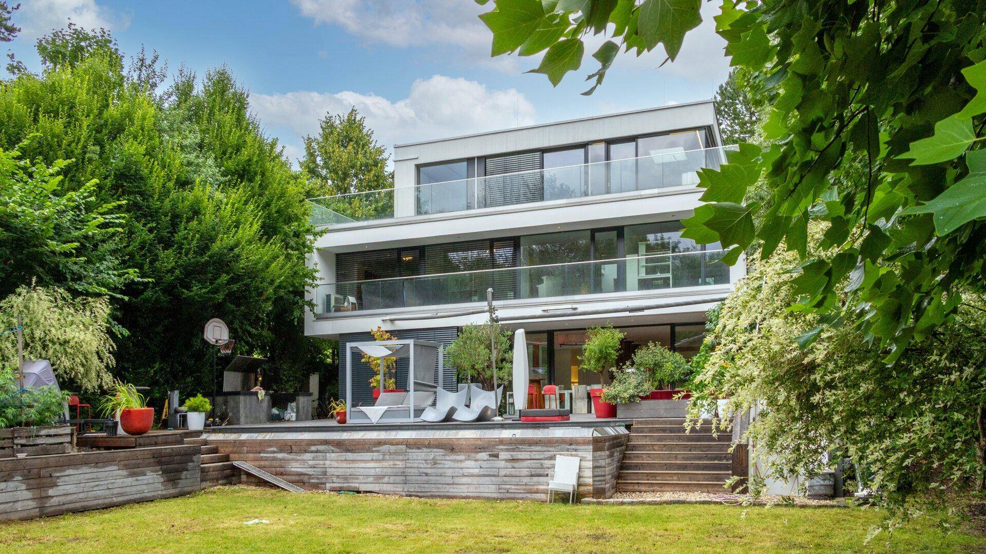 Moderne Villa mit weitläufigem Garten, Poolbereich und Basketballkorb, umgeben von viel Grün.