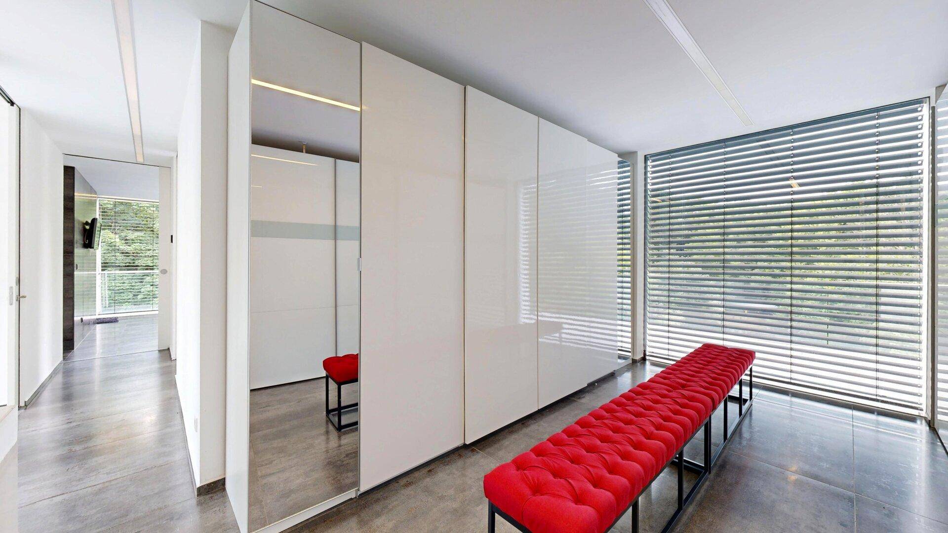 Moderner Flur mit glänzenden weißen Einbauschränken, großem Fenster und roter Polsterbank.