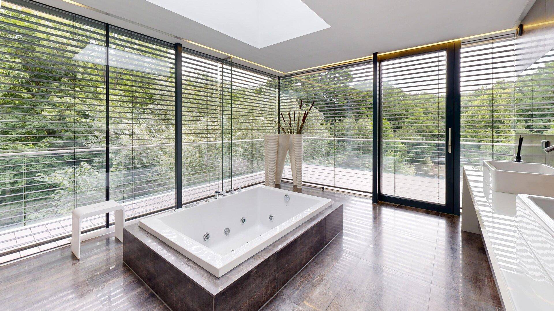 Geräumiges Badezimmer mit eingelassener Badewanne, großen Fenstern und Blick auf die grüne Umgebung.