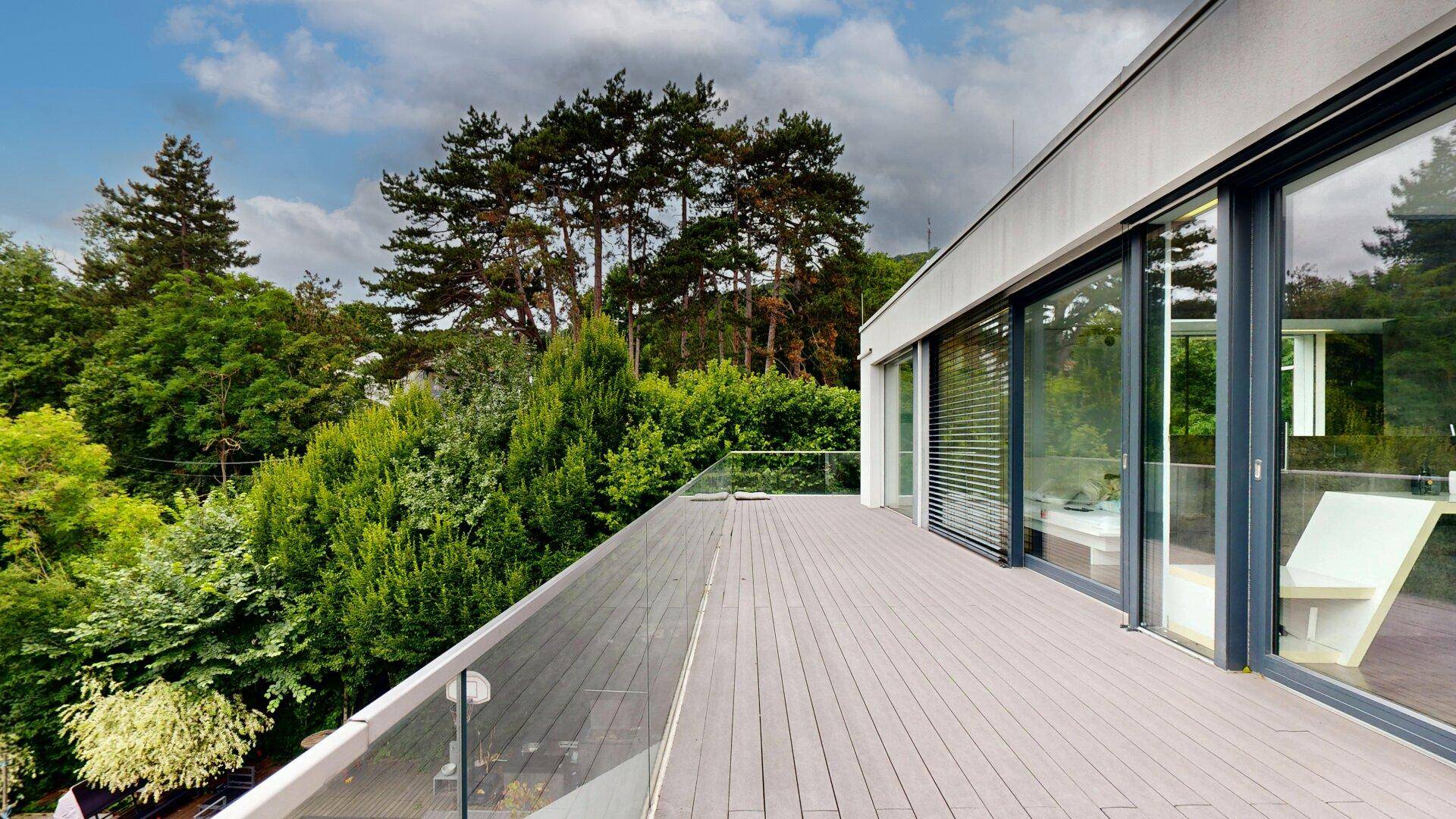 Weitläufige Terrasse mit Holzboden und Glasgeländer, umgeben von üppiger Natur und hohen Bäumen.