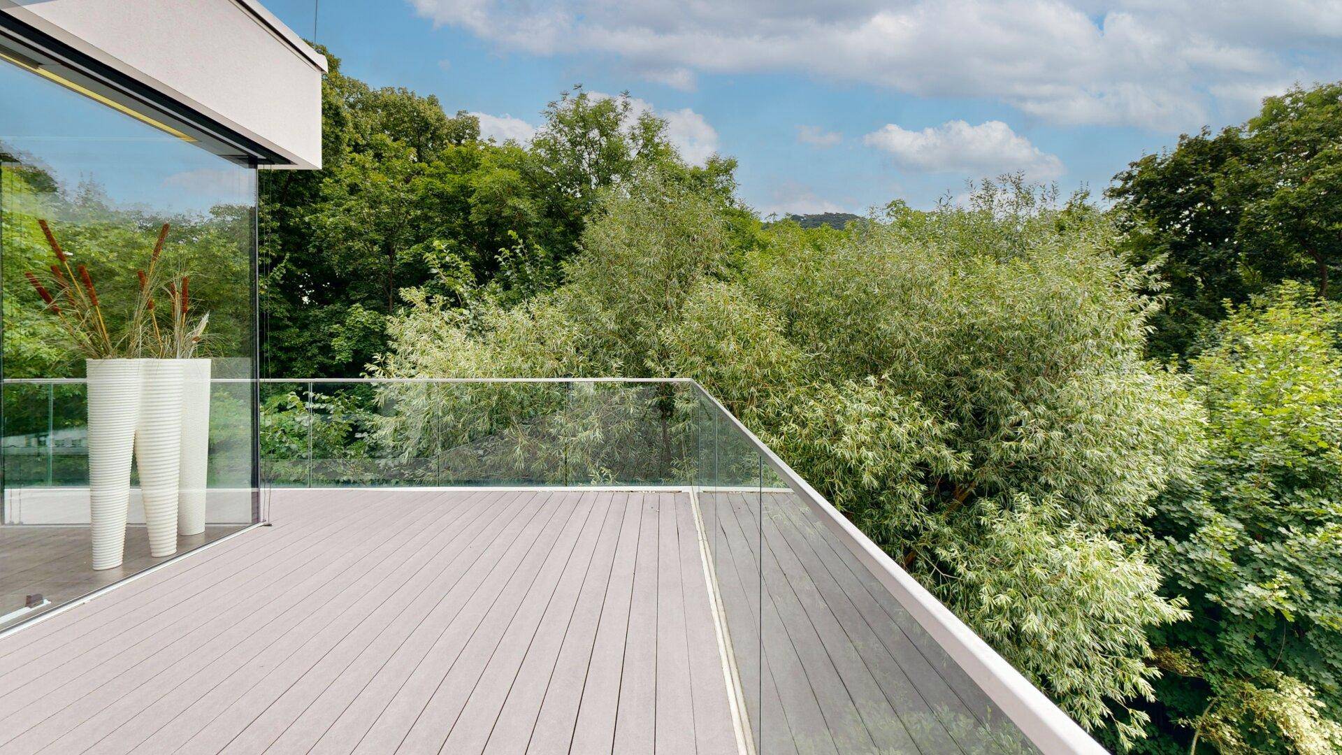 Helle Terrasse mit Glasgeländer und weitem Blick auf die umliegende grüne Landschaft und Bäume.
