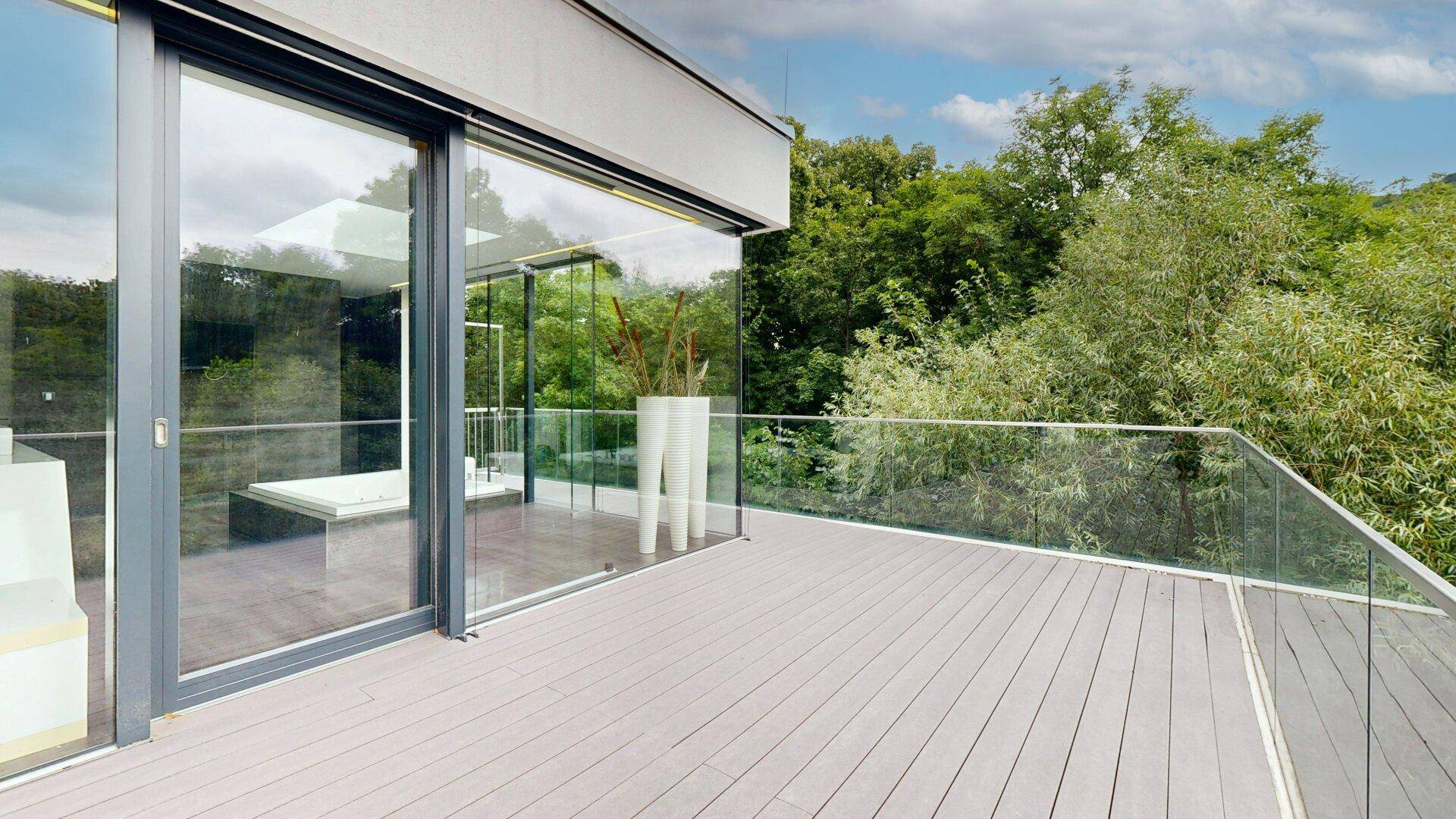 Große Terrasse mit Holzboden und Glasgeländer, bietet einen weiten Blick auf die grüne Umgebung.