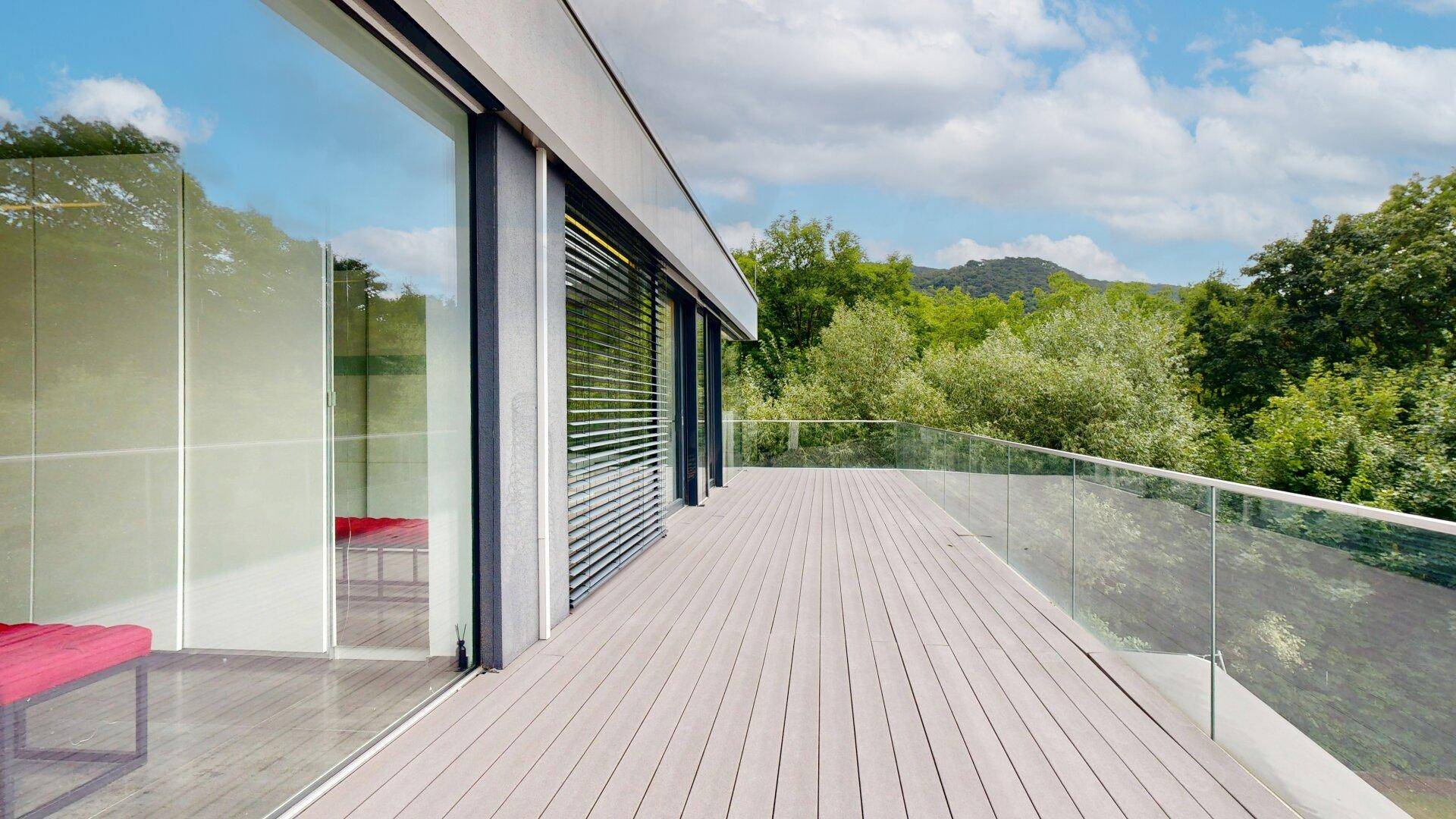 Helle Terrasse mit Holzboden und Glasgeländer, bietet einen weiten Blick auf die grüne Hügellandschaft.