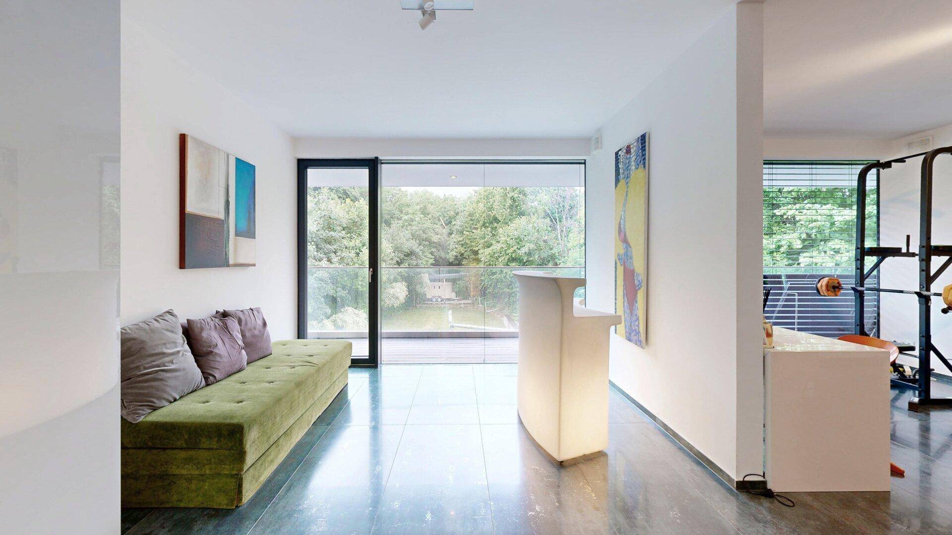 Modernes Wohnzimmer mit grüner Couch, Bar und Zugang zum Balkon mit Blick ins Grüne.