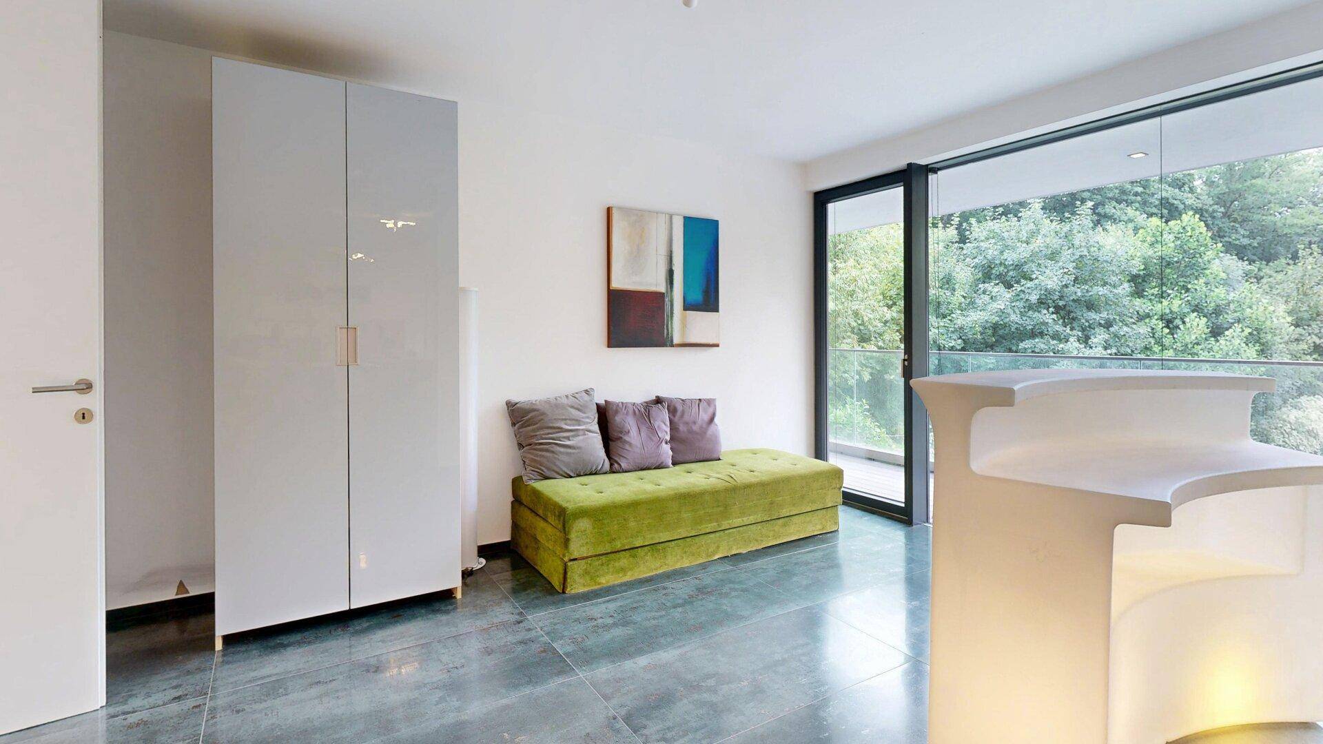 Helles Zimmer mit grüner Sitzbank, Schrank und Zugang zu einem Balkon mit viel Grün.